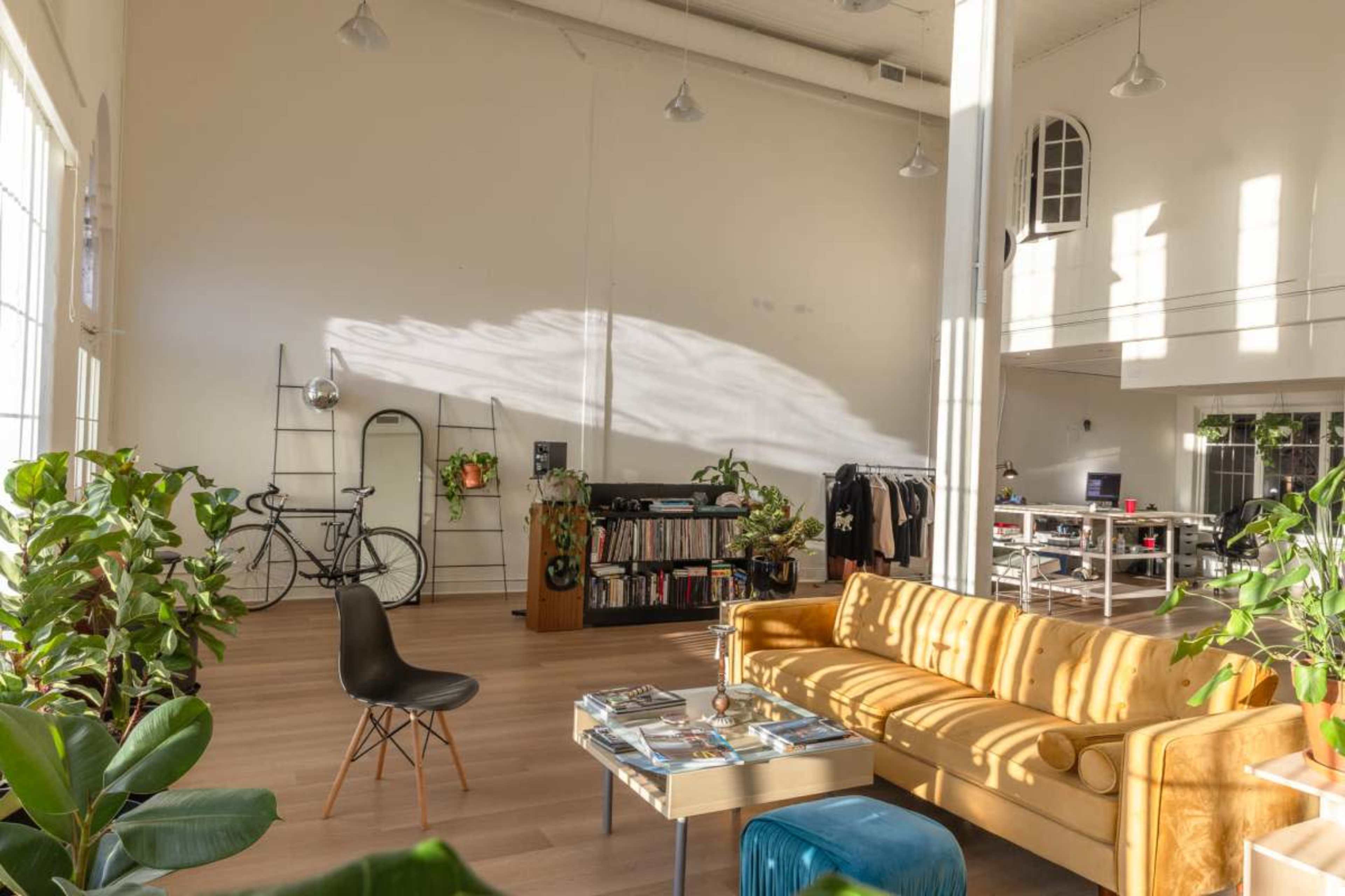 The image shows a spacious room with large windows, a yellow sofa, a coffee table filled with magazines, and various plants, along with a bicycle and shelves in the background.