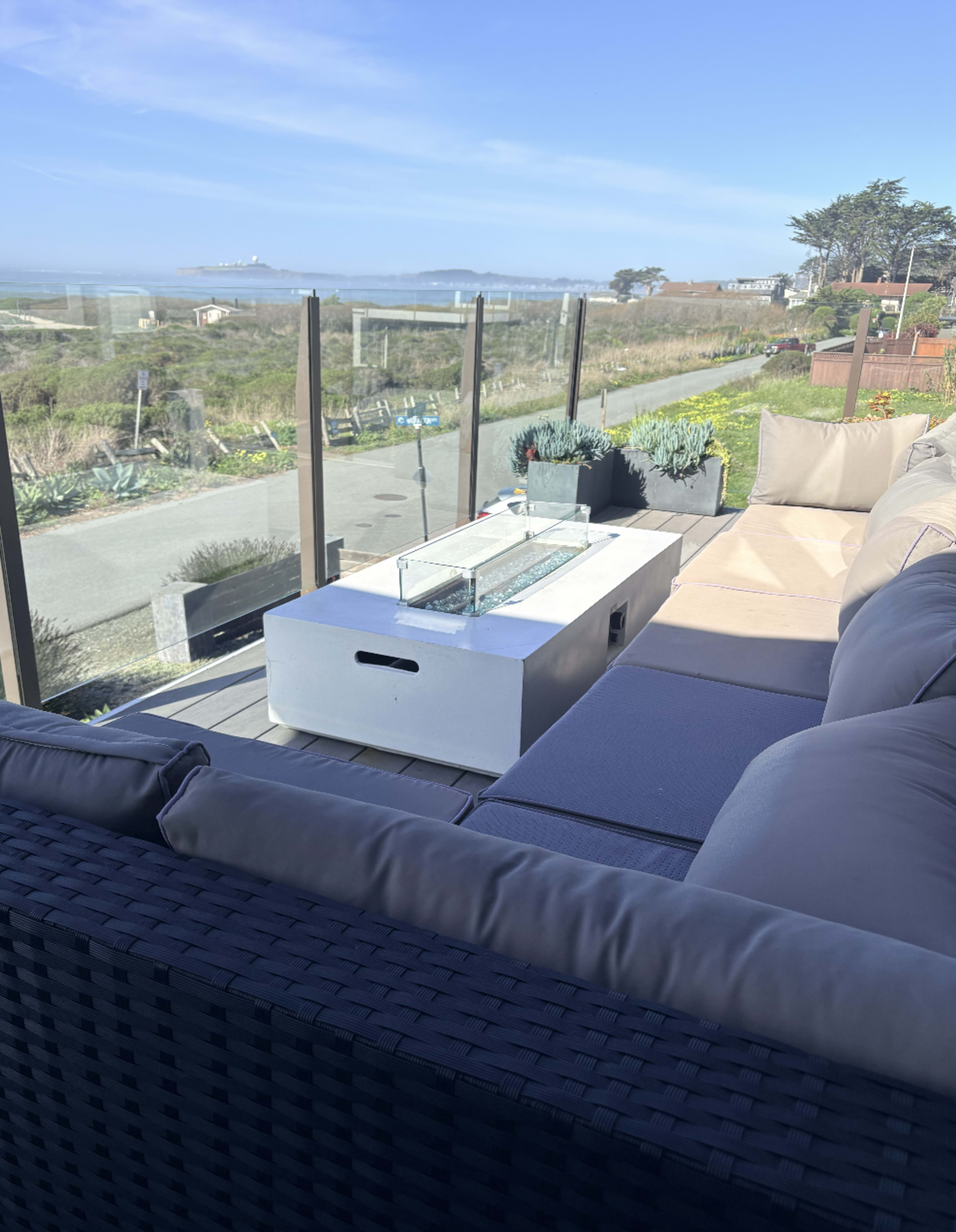 A modern outdoor seating area features a glass railing, a fire table, and views of a coastal landscape.