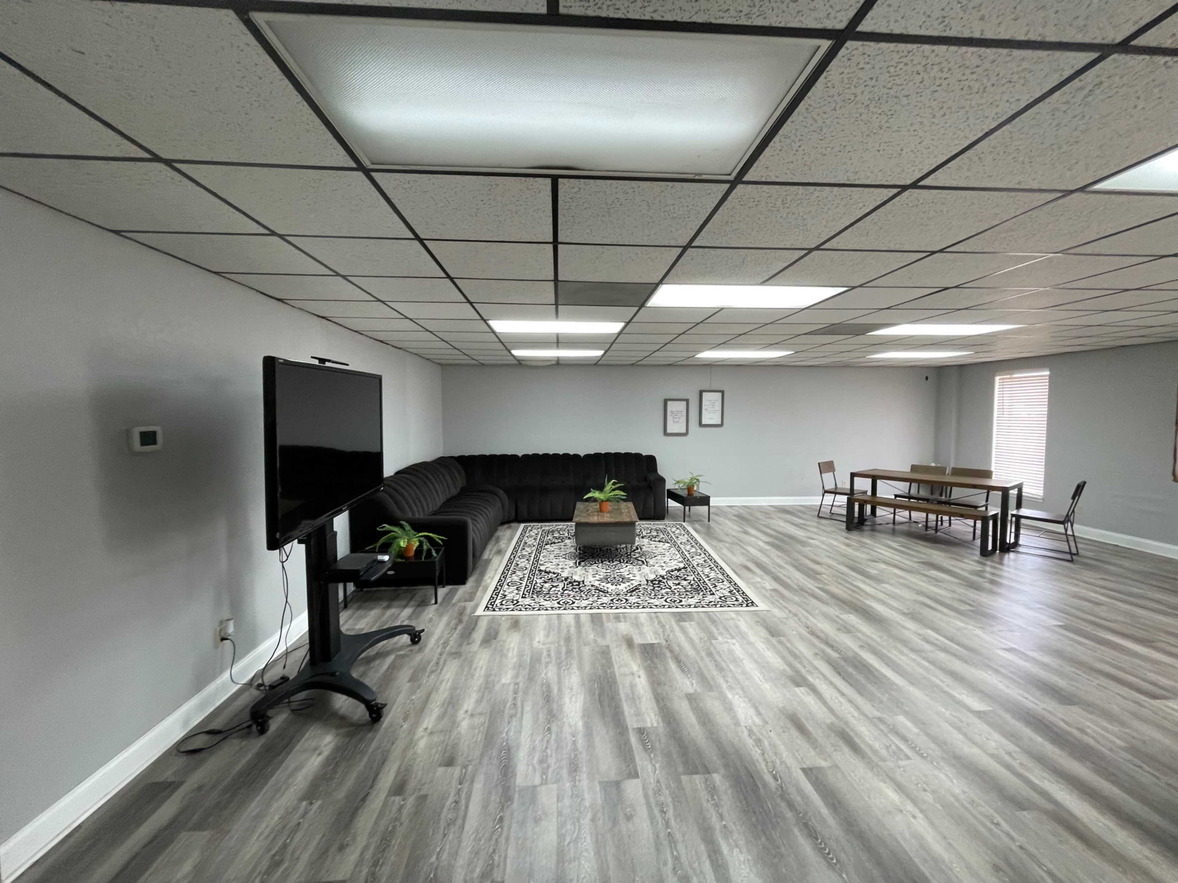 The image shows a spacious room with gray walls, a dark sectional couch, a decorative rug, a television on a stand, and a wooden dining table with chairs.