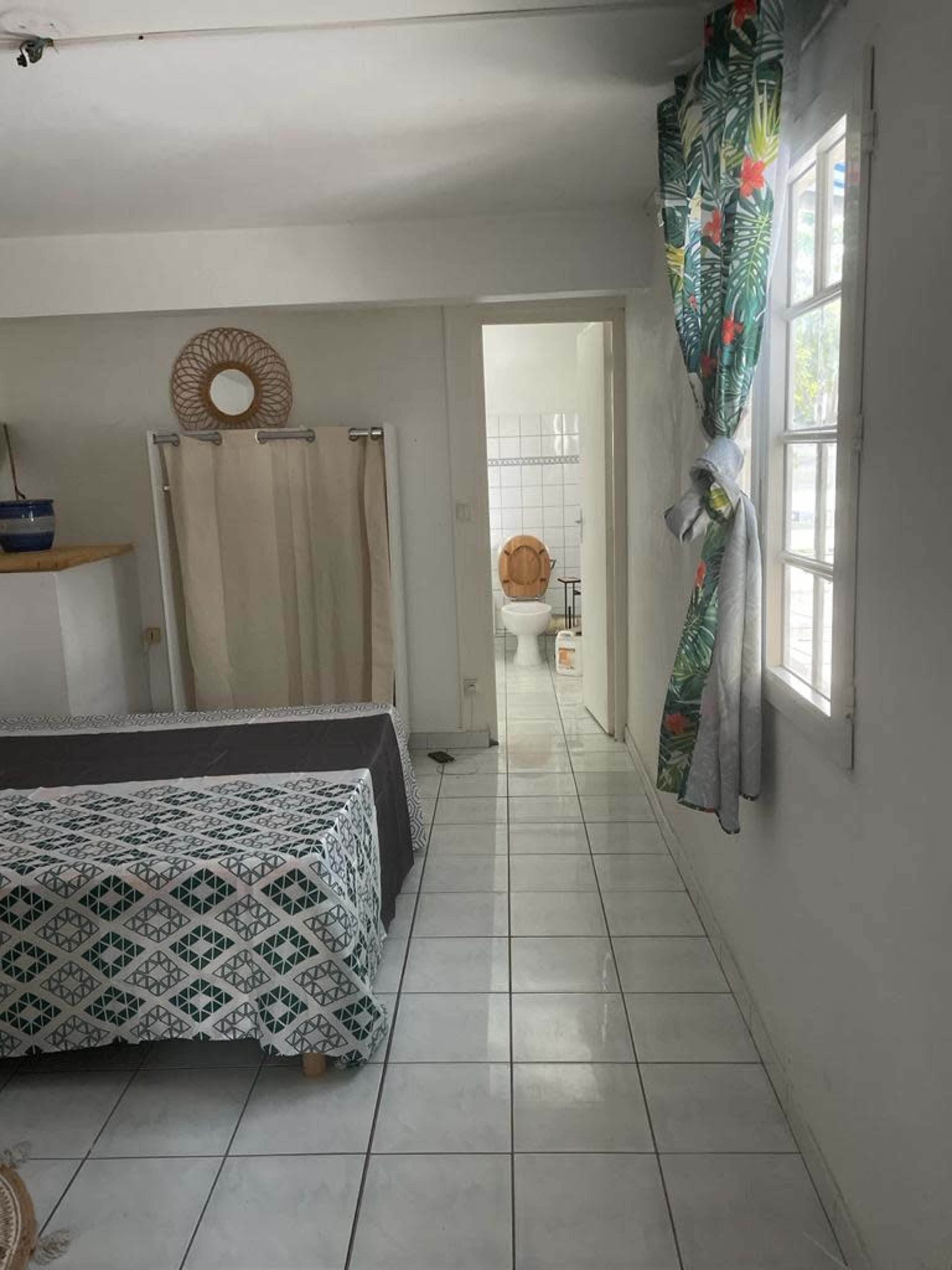 A simple room with a bed, a window draped with floral curtains, and a bathroom visible in the background.