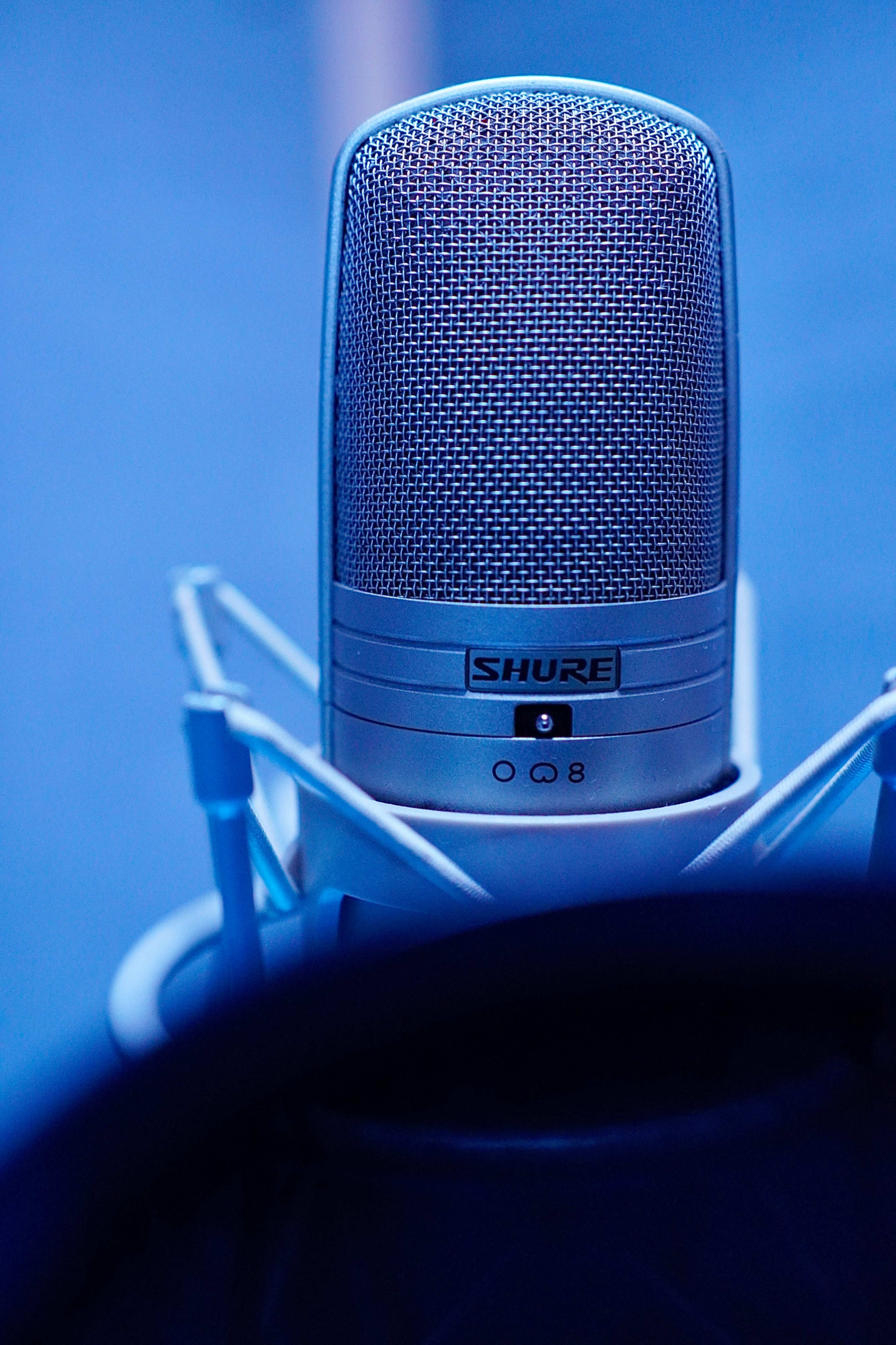 A silver microphone is positioned against a blue background, mounted on a white shock mount.