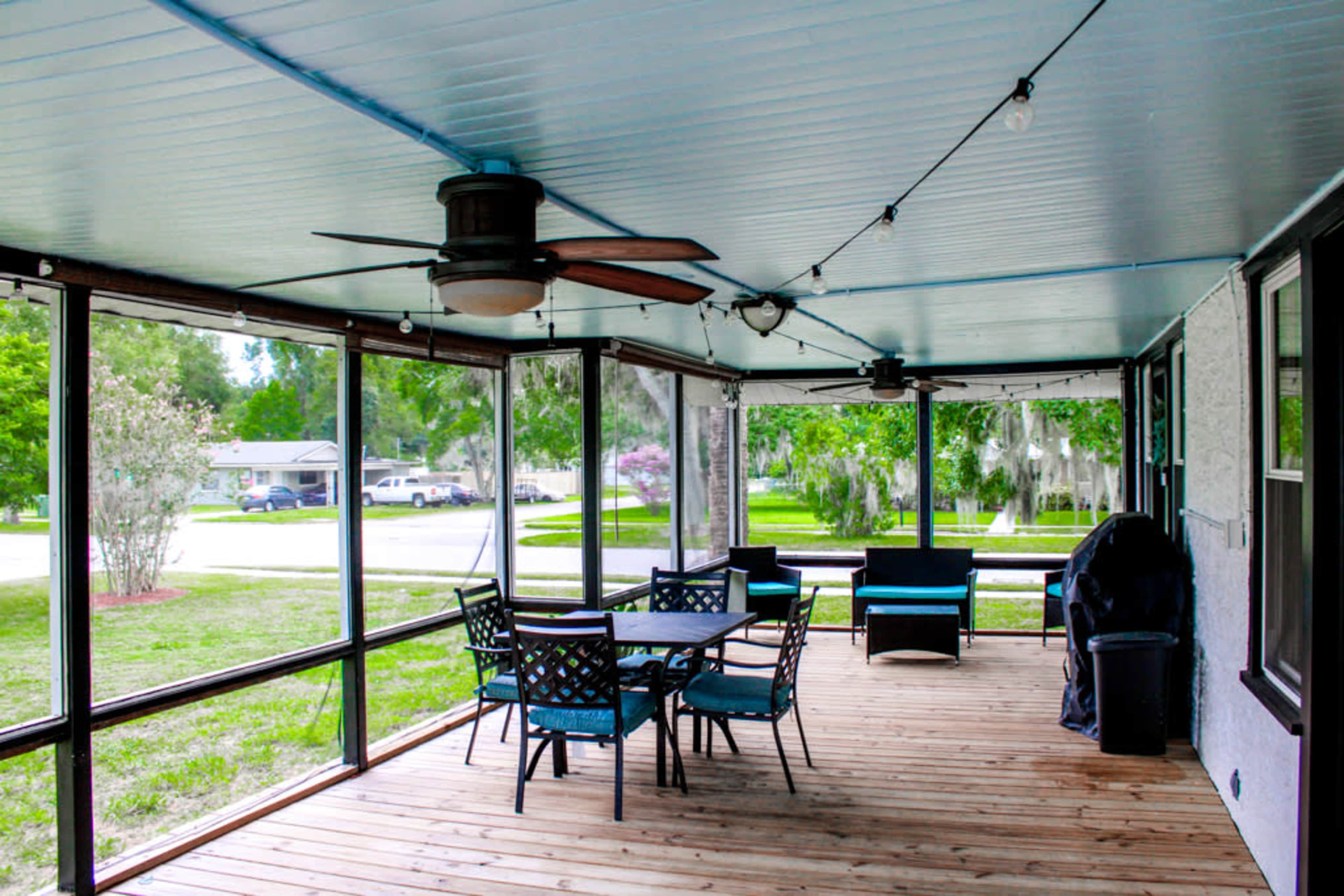 A screened porch features a wooden floor, table with chairs, and a ceiling fan, surrounded by greenery.