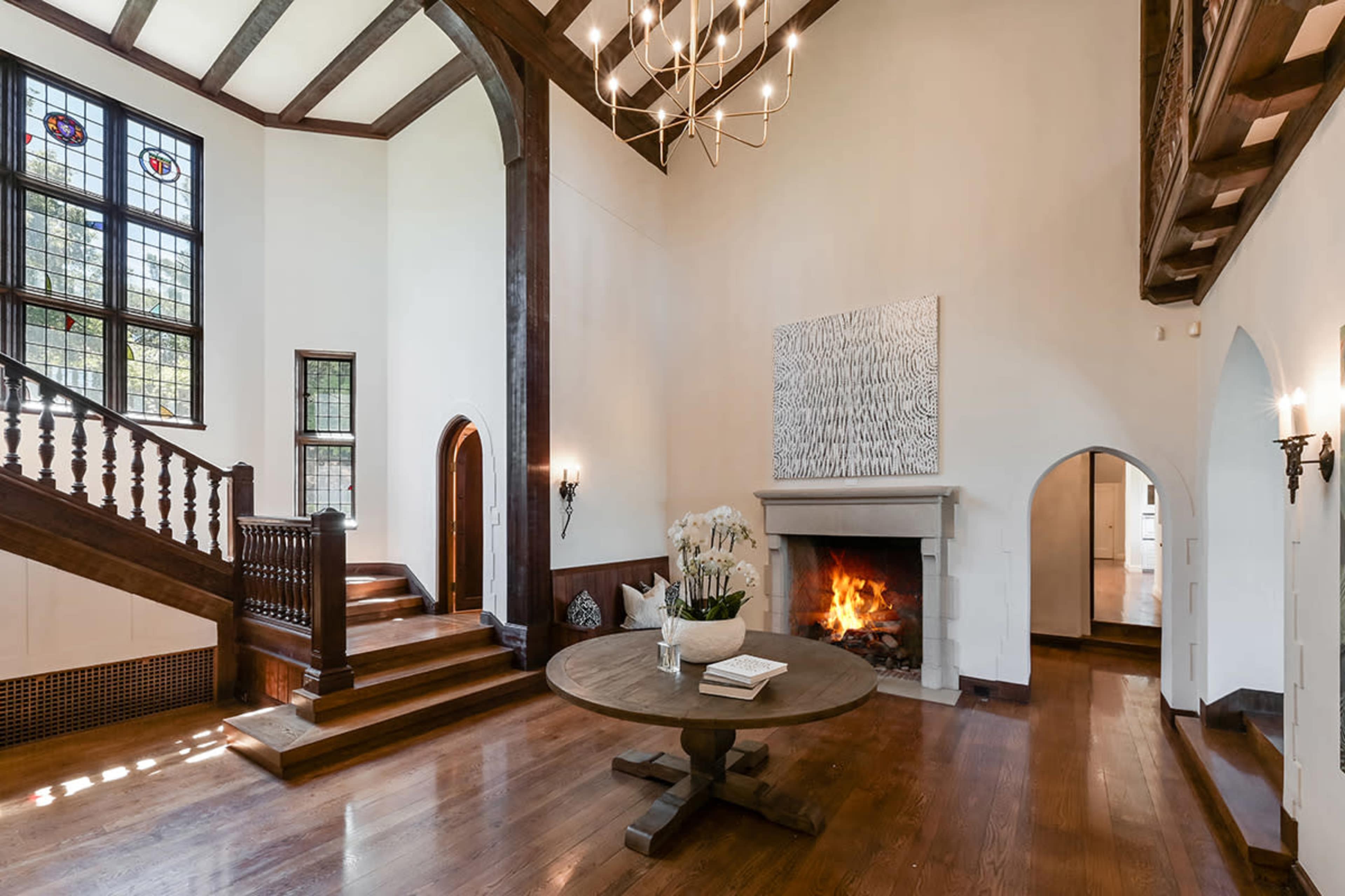 The image shows a spacious living area with a large stone fireplace, dark wooden beams, and an elegant staircase leading to a second level.