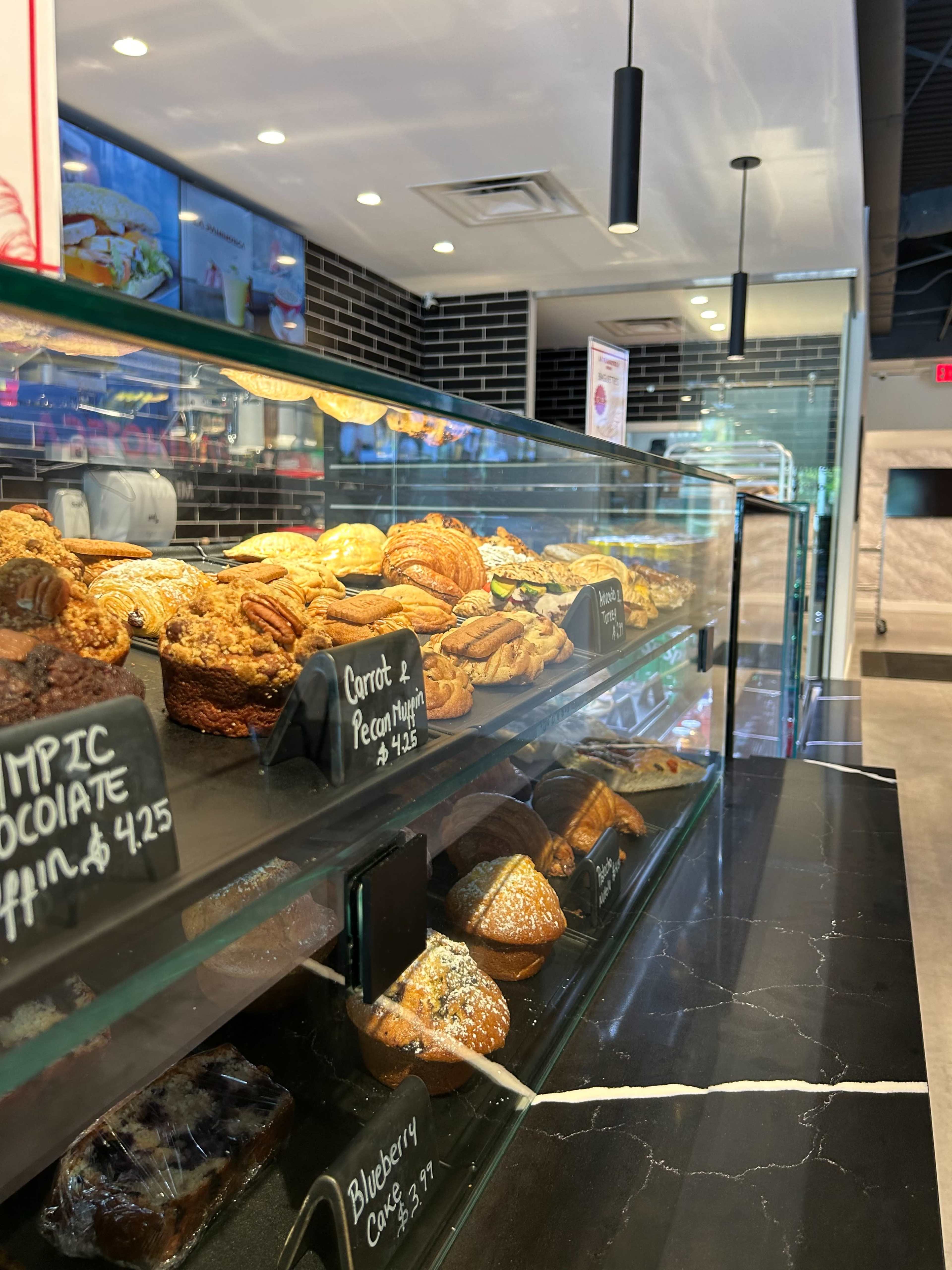 The image shows a display case in a bakery filled with various pastries, including muffins and cookies, arranged on shelves with labels indicating their prices.