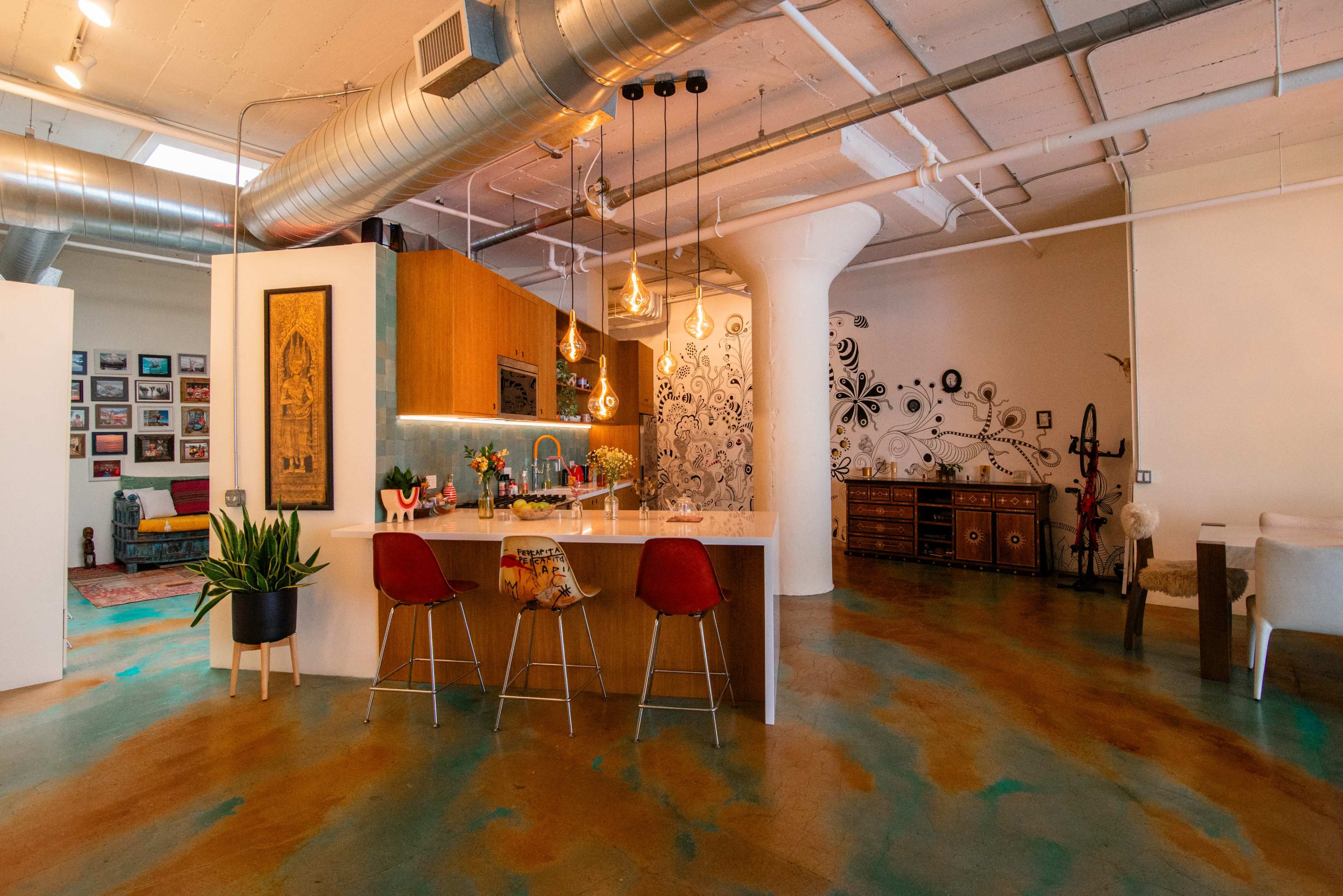 The image shows a modern industrial-style kitchen and living area with a bar counter, colorful floors, and eclectic decor elements.