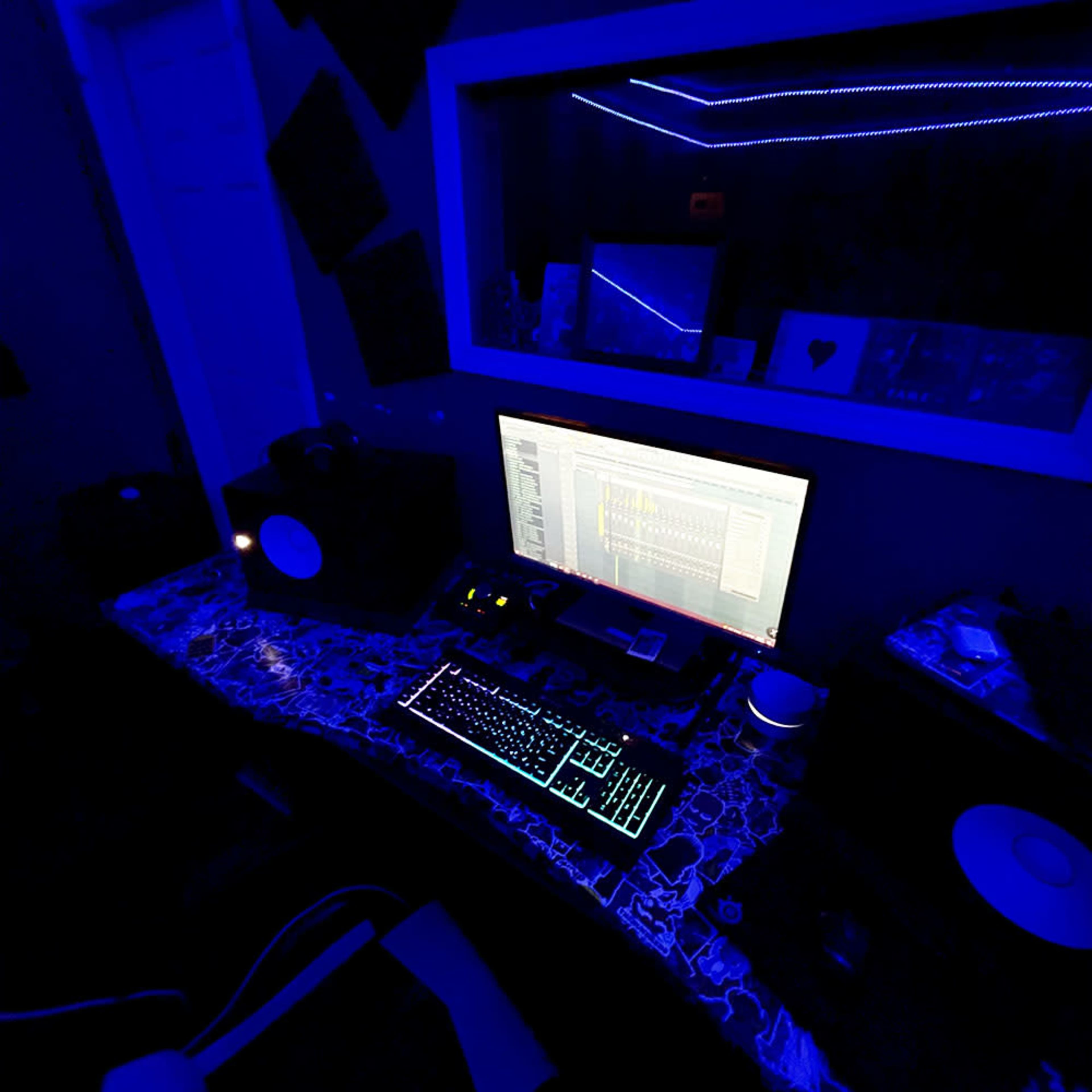 A dimly lit music production workspace features a computer monitor, illuminated keyboard, and speakers on a patterned desk.