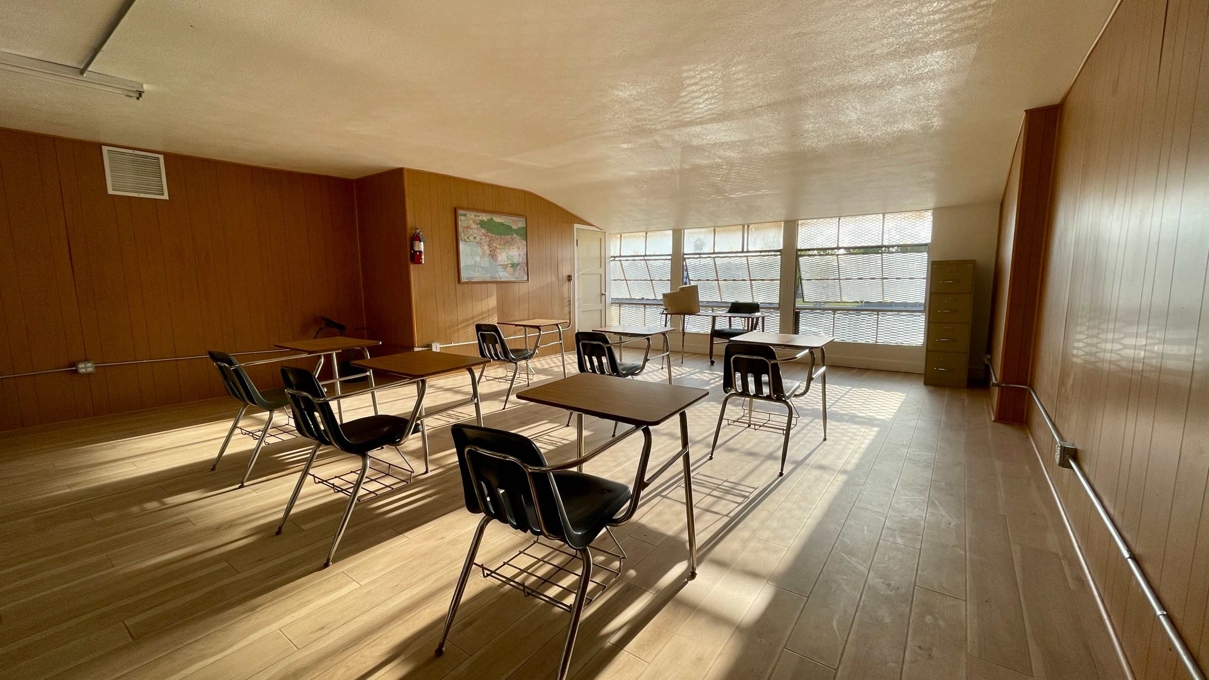A sparsely furnished classroom with several desks arranged in rows and a large window allowing natural light to fill the space.