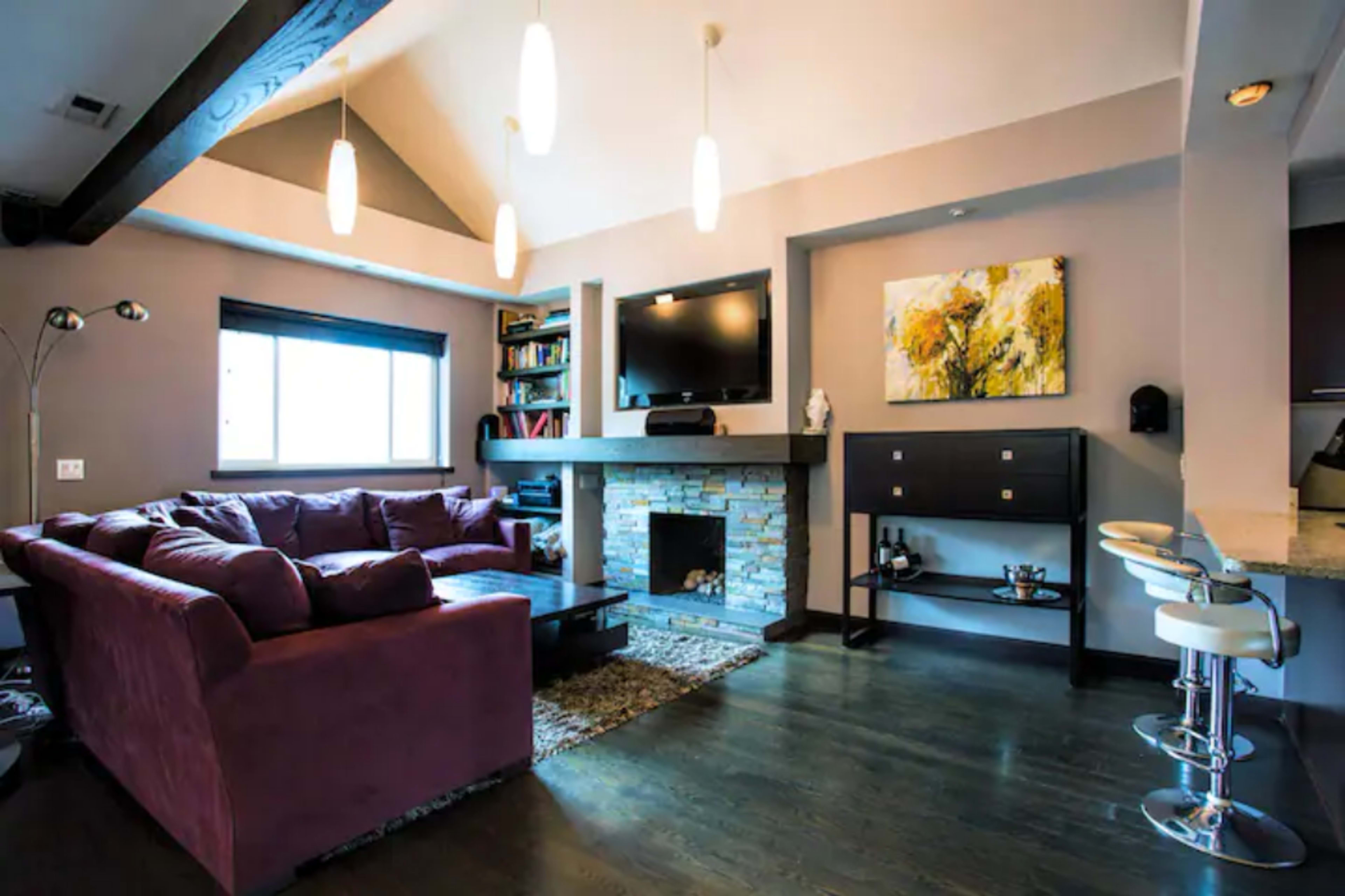 The living room features a purple sectional sofa facing a stone fireplace with a mounted television above it, accompanied by a bookshelf and a small dining area with bar stools.