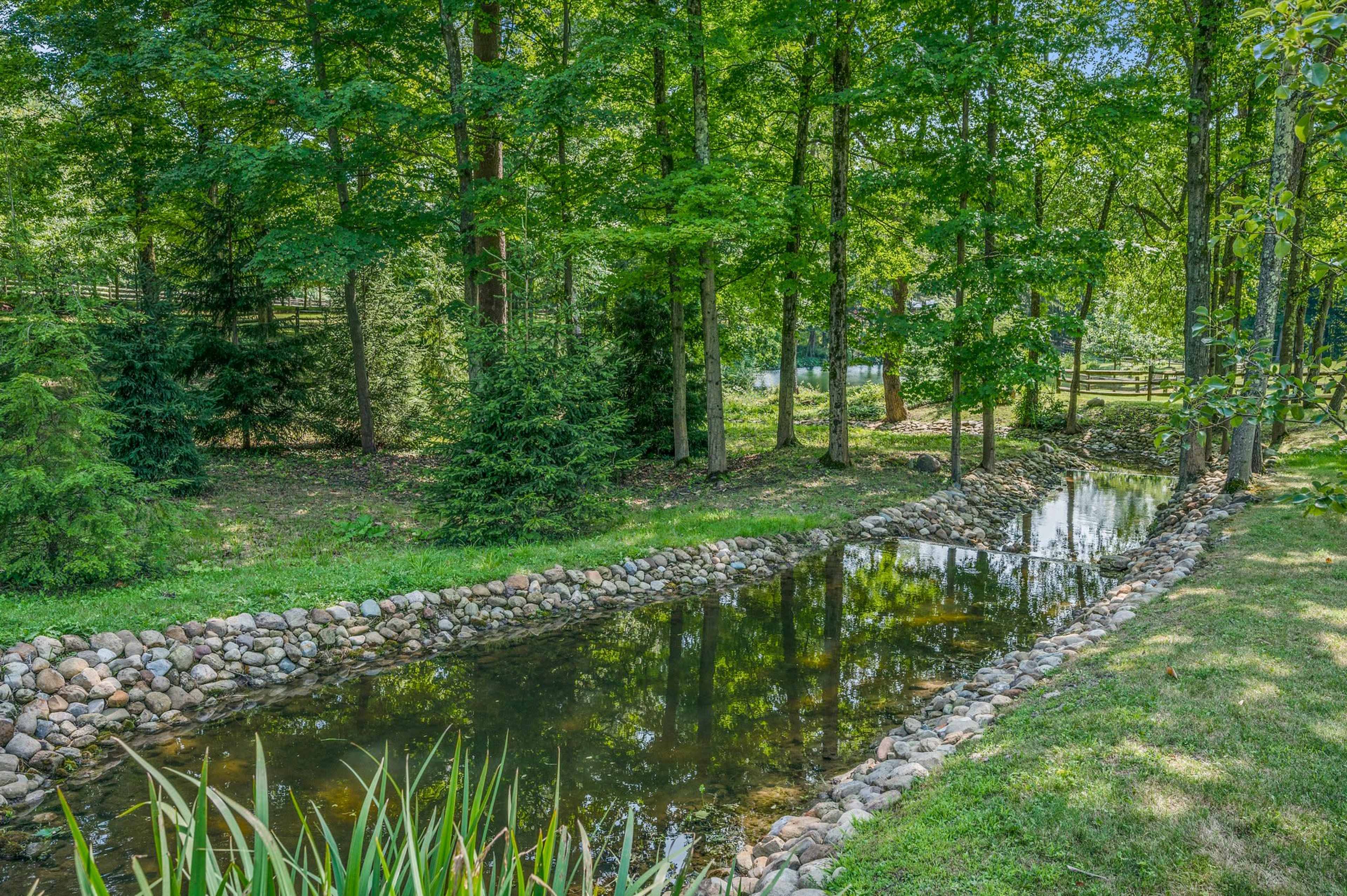 The image shows a serene landscape featuring a narrow, water-filled channel bordered by stones, surrounded by lush green trees and shrubs.