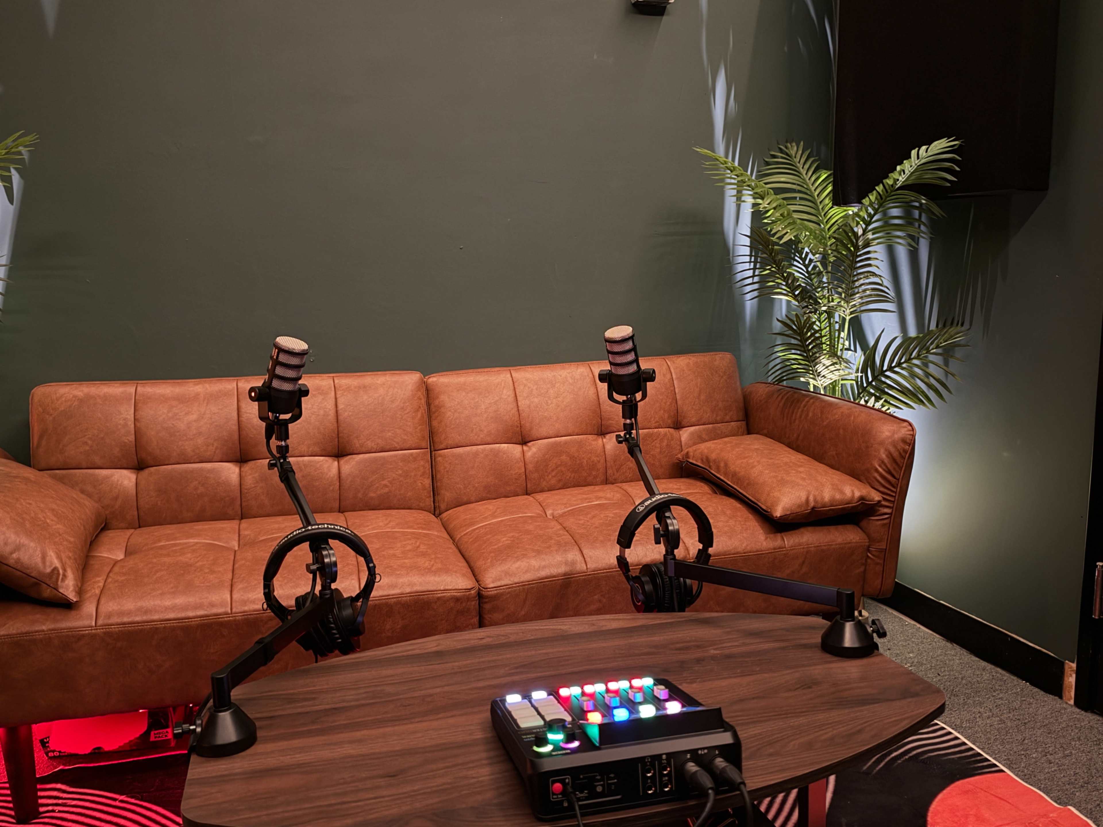 The image shows a modern podcast recording setup featuring a brown leather couch, two microphones on adjustable stands, and a control panel with colorful buttons on a wooden table.