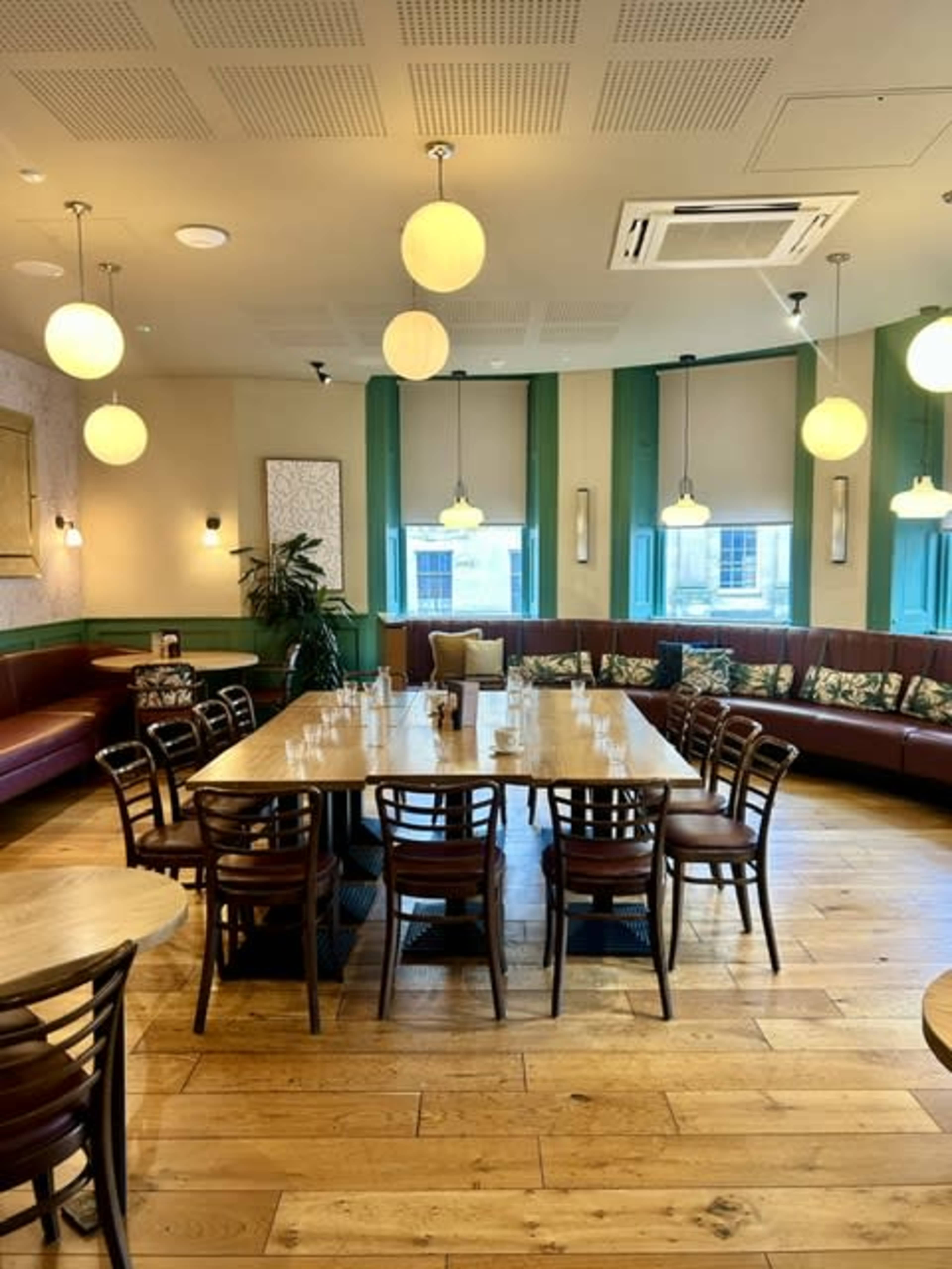 The image shows a modern dining area with wooden tables and chairs, featuring a large round table at the center and a cozy booth seating arrangement along the walls.
