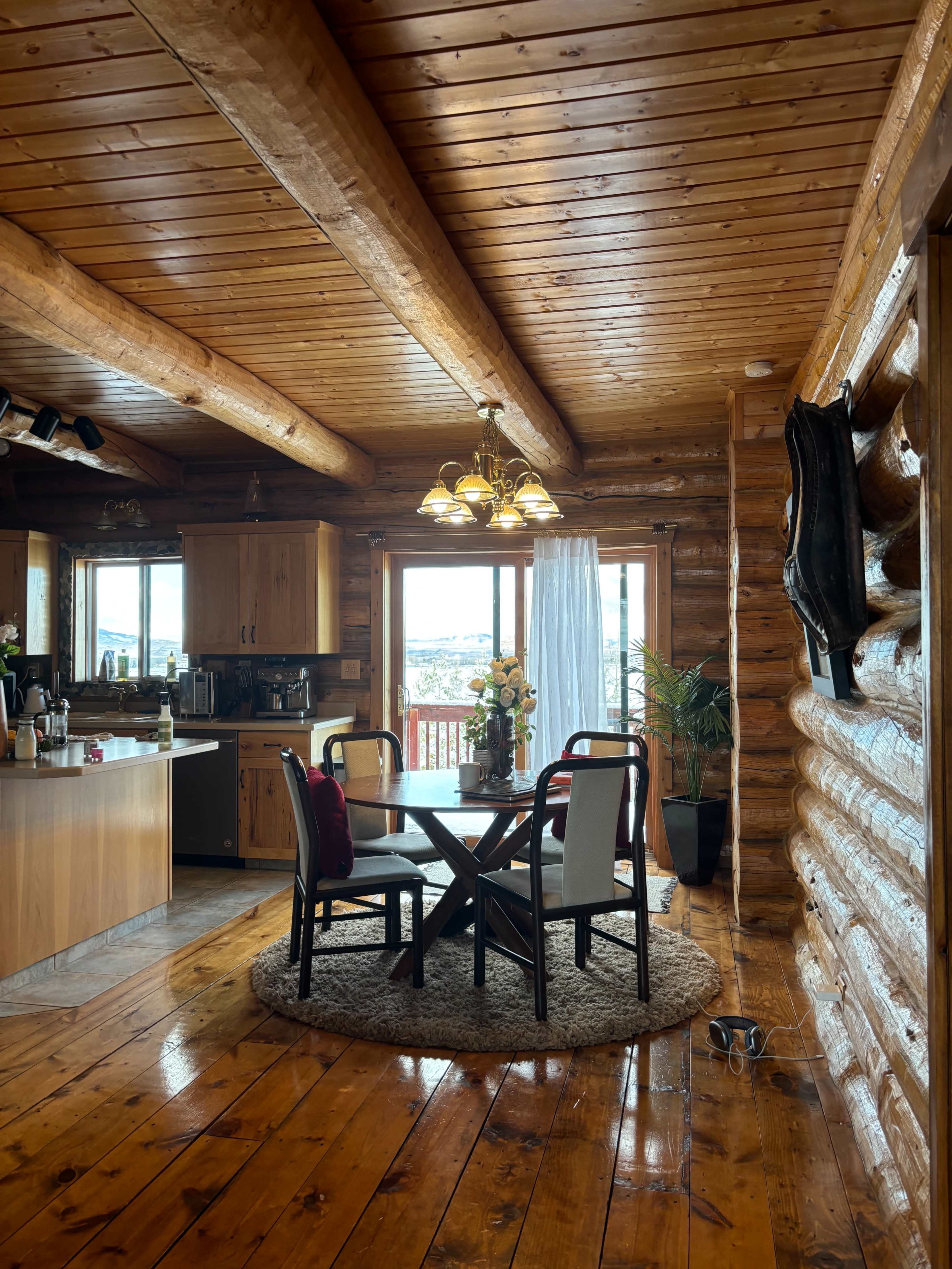 The image shows a cozy log cabin interior featuring a dining area with a round table, four chairs, and a view of the outdoor scenery through large windows.