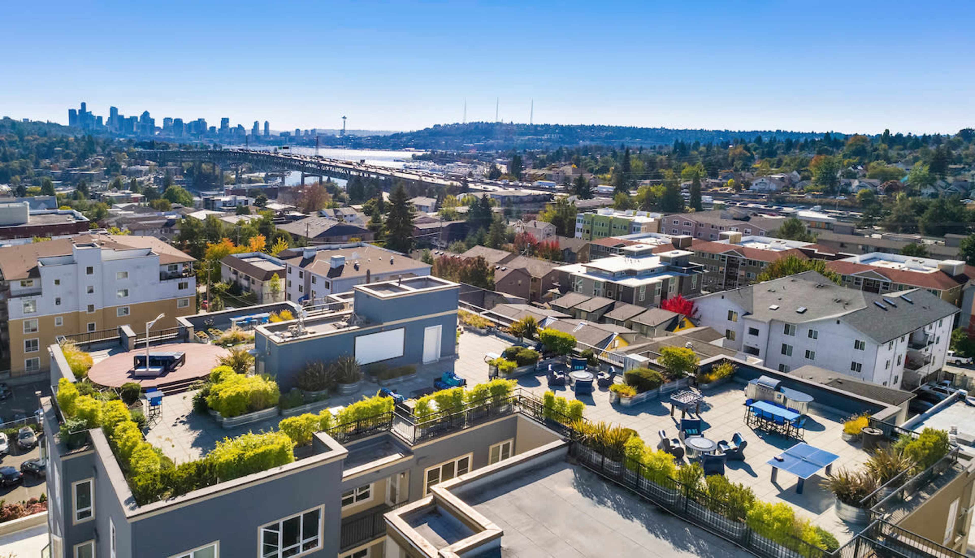 An aerial view of a residential area with modern buildings and a rooftop deck, overlooking a city skyline and a bridge across a body of water.