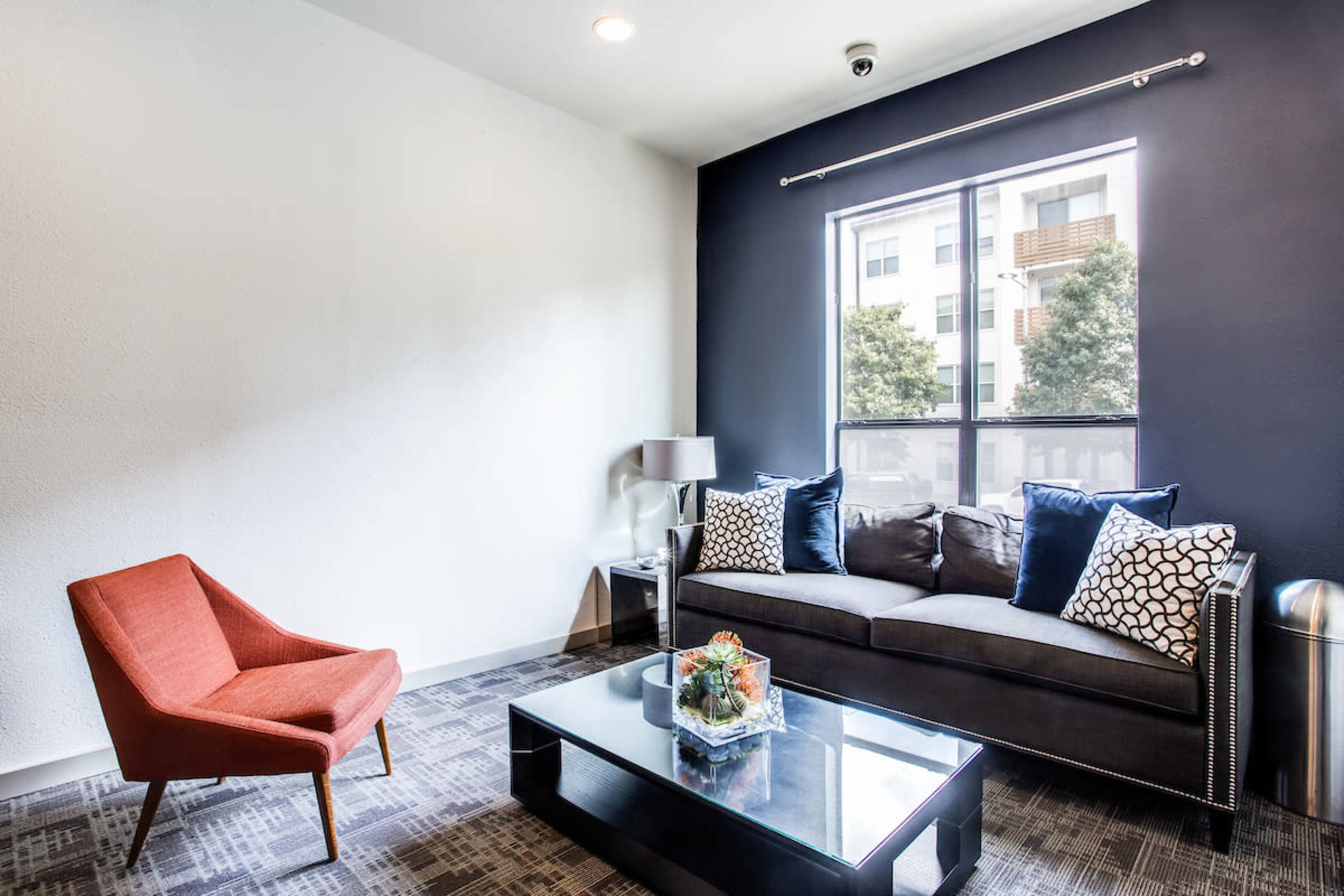 A modern lounge area features a dark sofa with patterned pillows, an orange armchair, a glass coffee table, and a window overlooking an exterior view.