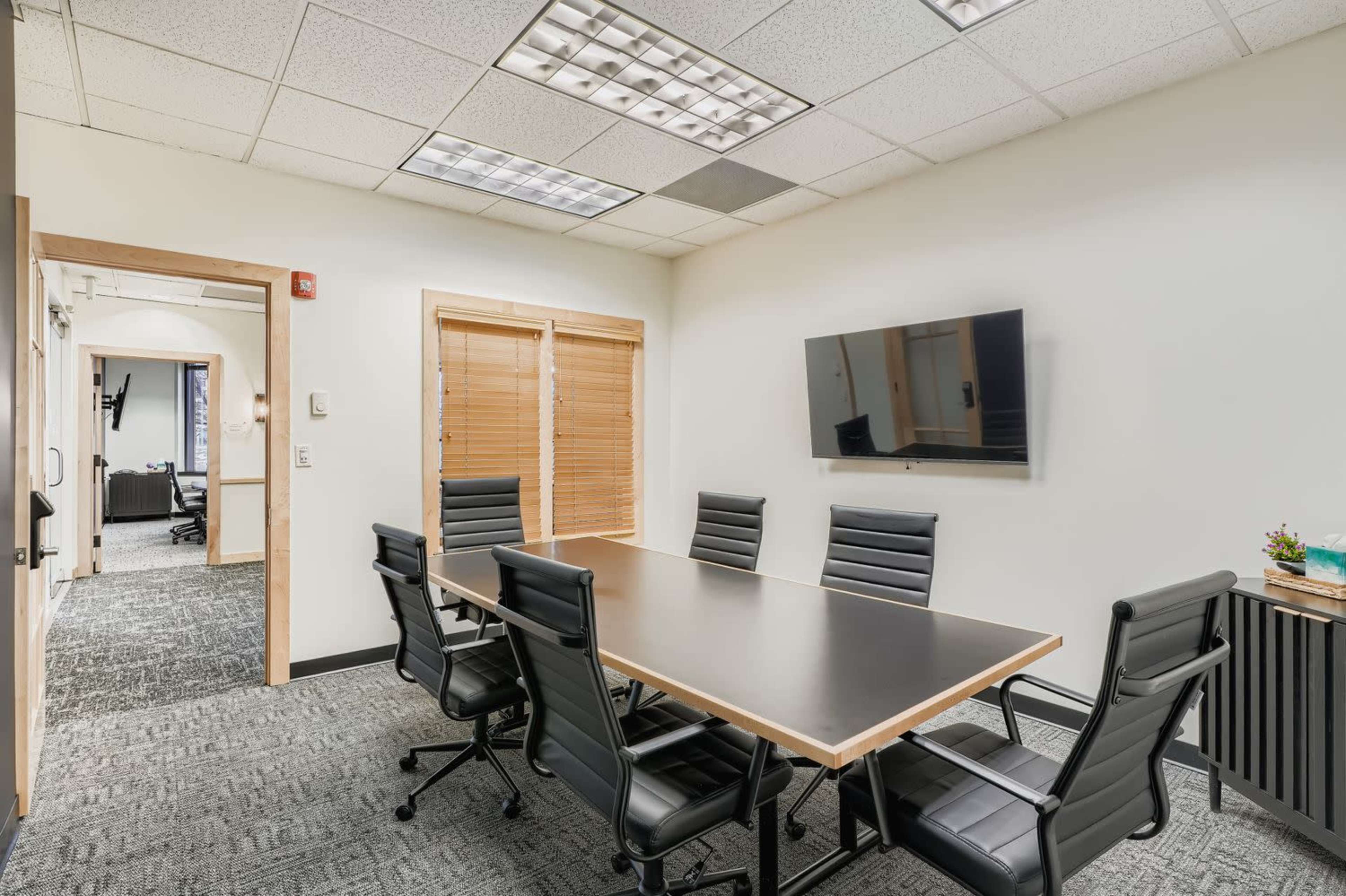 The image shows a conference room with a long black table surrounded by six black leather chairs, and a wall-mounted television.
