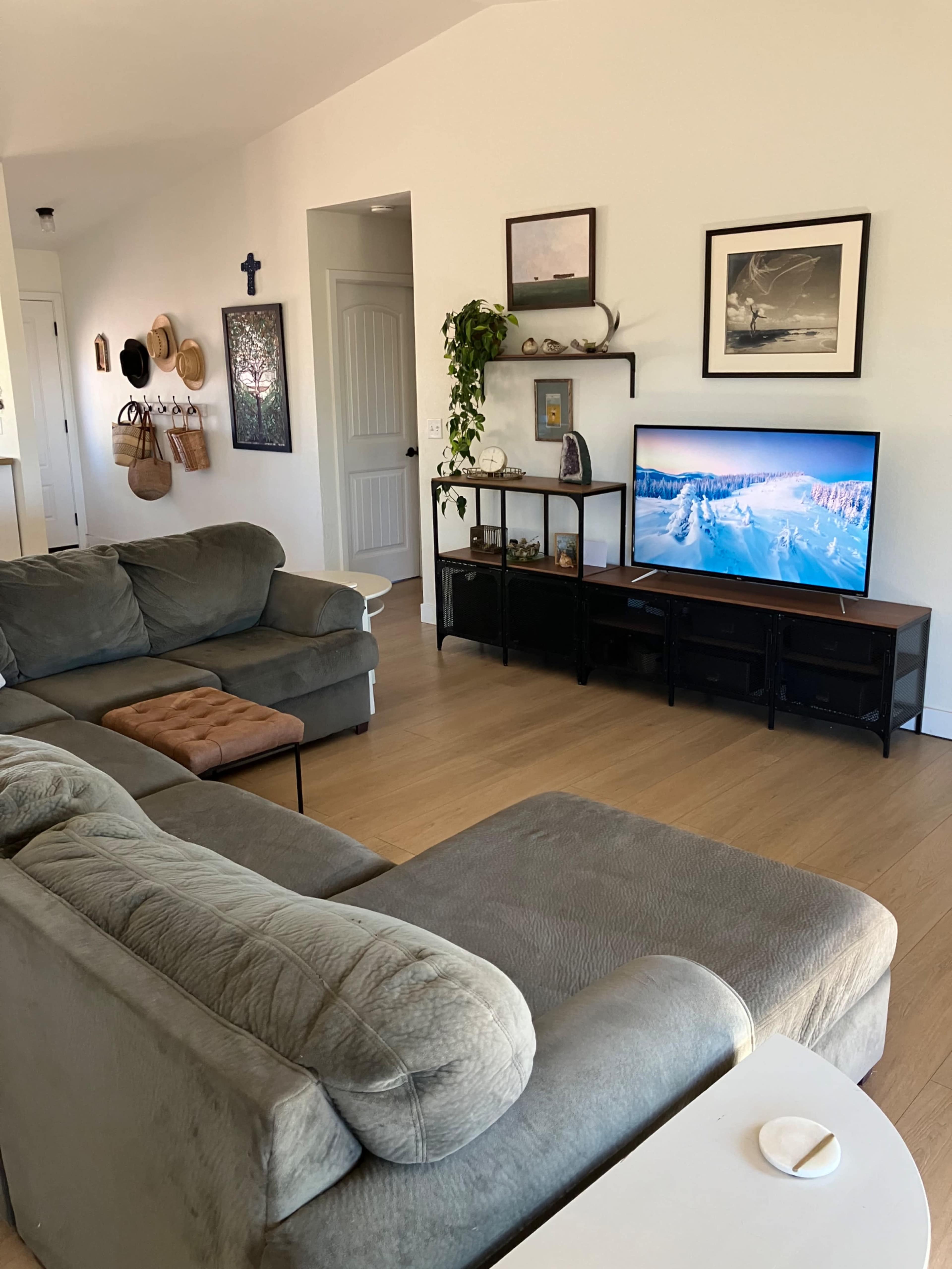 The living room features a gray sectional sofa, a television on a black stand displaying a mountain landscape, and various decor items on the walls and shelves.