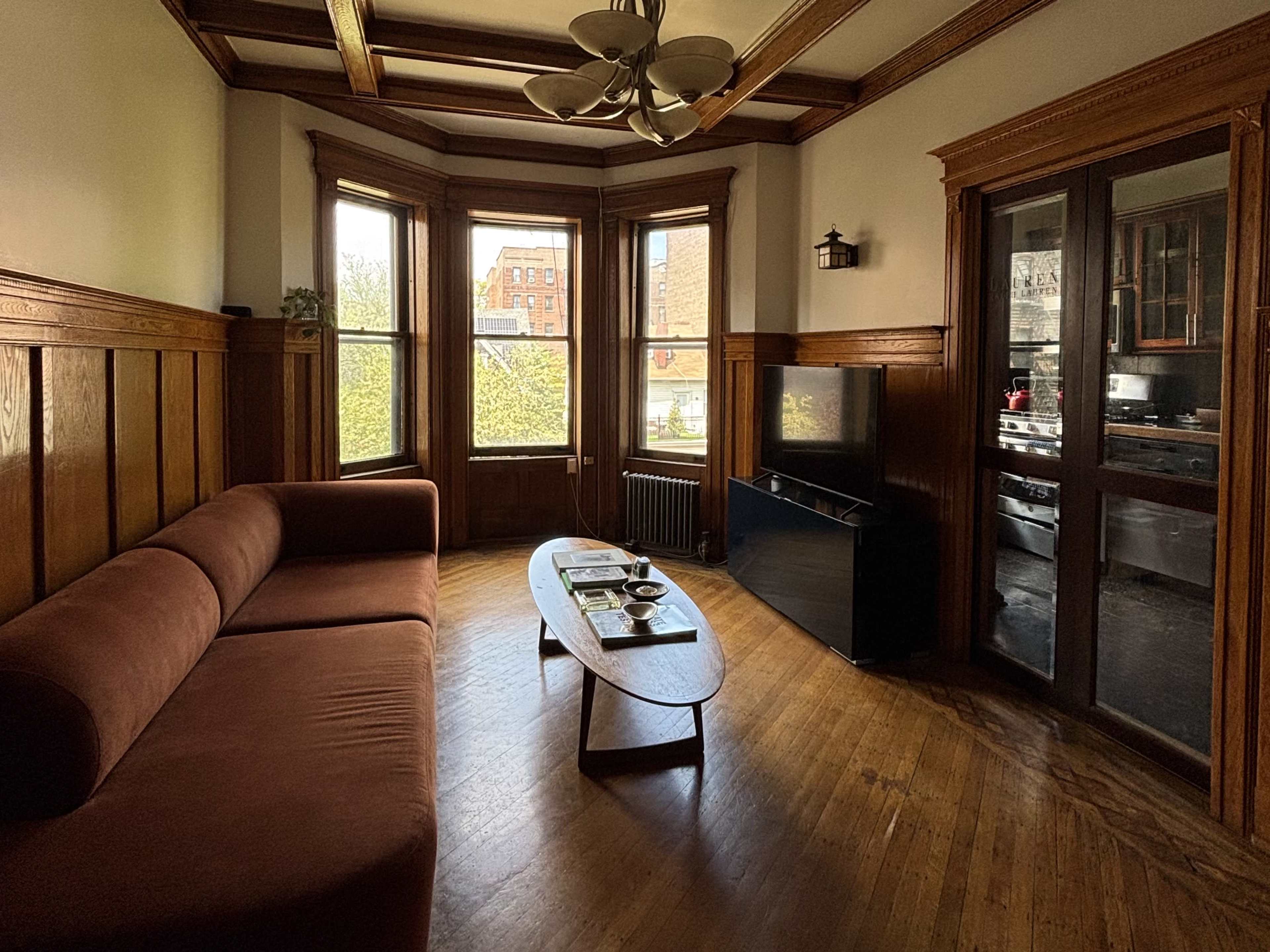 The image depicts a furnished living room with a brown sofa, a coffee table, and large windows allowing natural light to illuminate the wooden paneling and flooring.