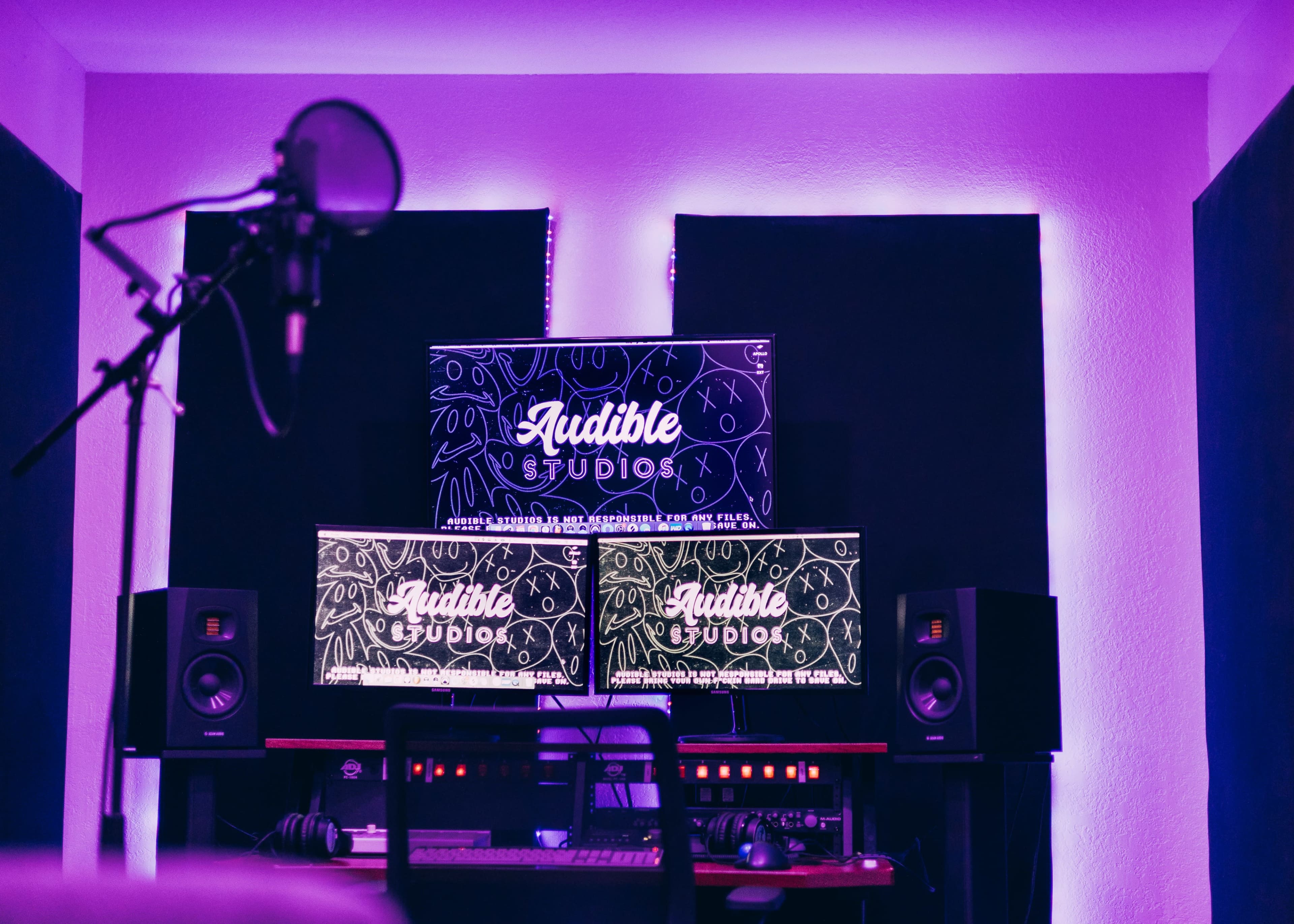 A modern music studio with two monitors displaying the logo "Audible Studios" and a microphone on a stand in the foreground.