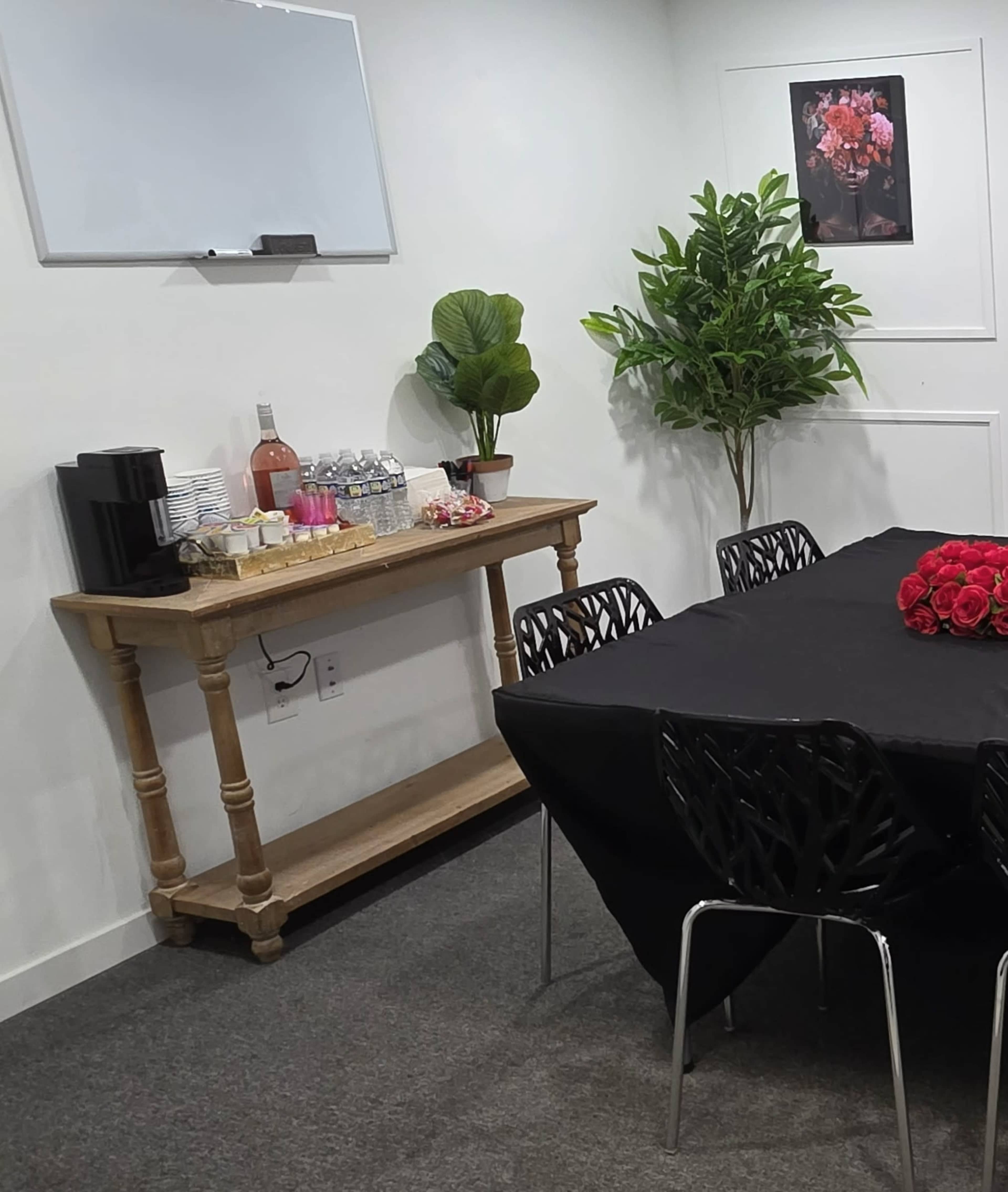 The image shows a small meeting room with a black table set for an event, featuring a wooden side table with refreshments, a potted plant, and a whiteboard on the wall.