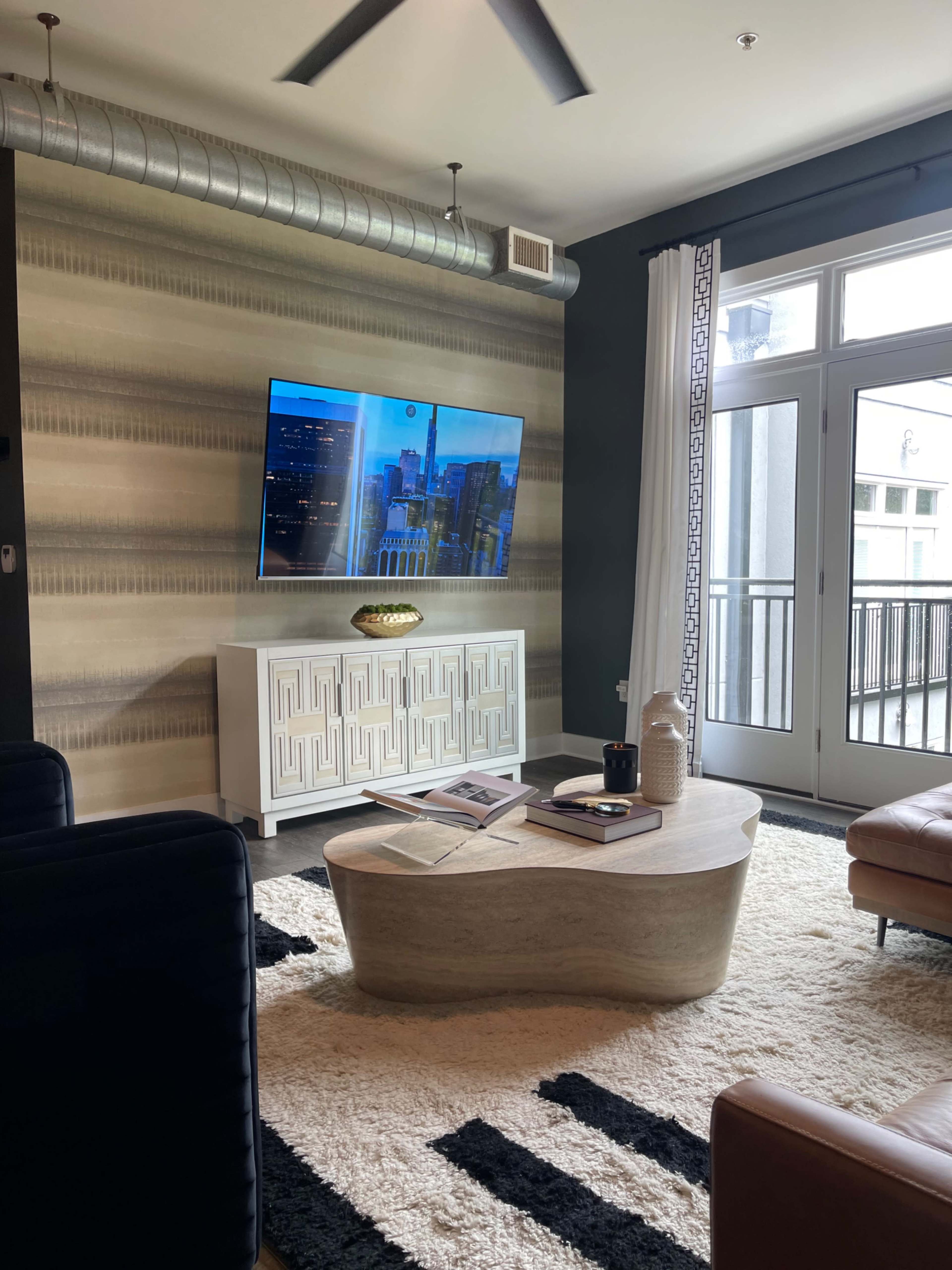 A modern living room features a television mounted on a patterned wall, a low coffee table, and comfortable seating with large windows that open to a balcony.