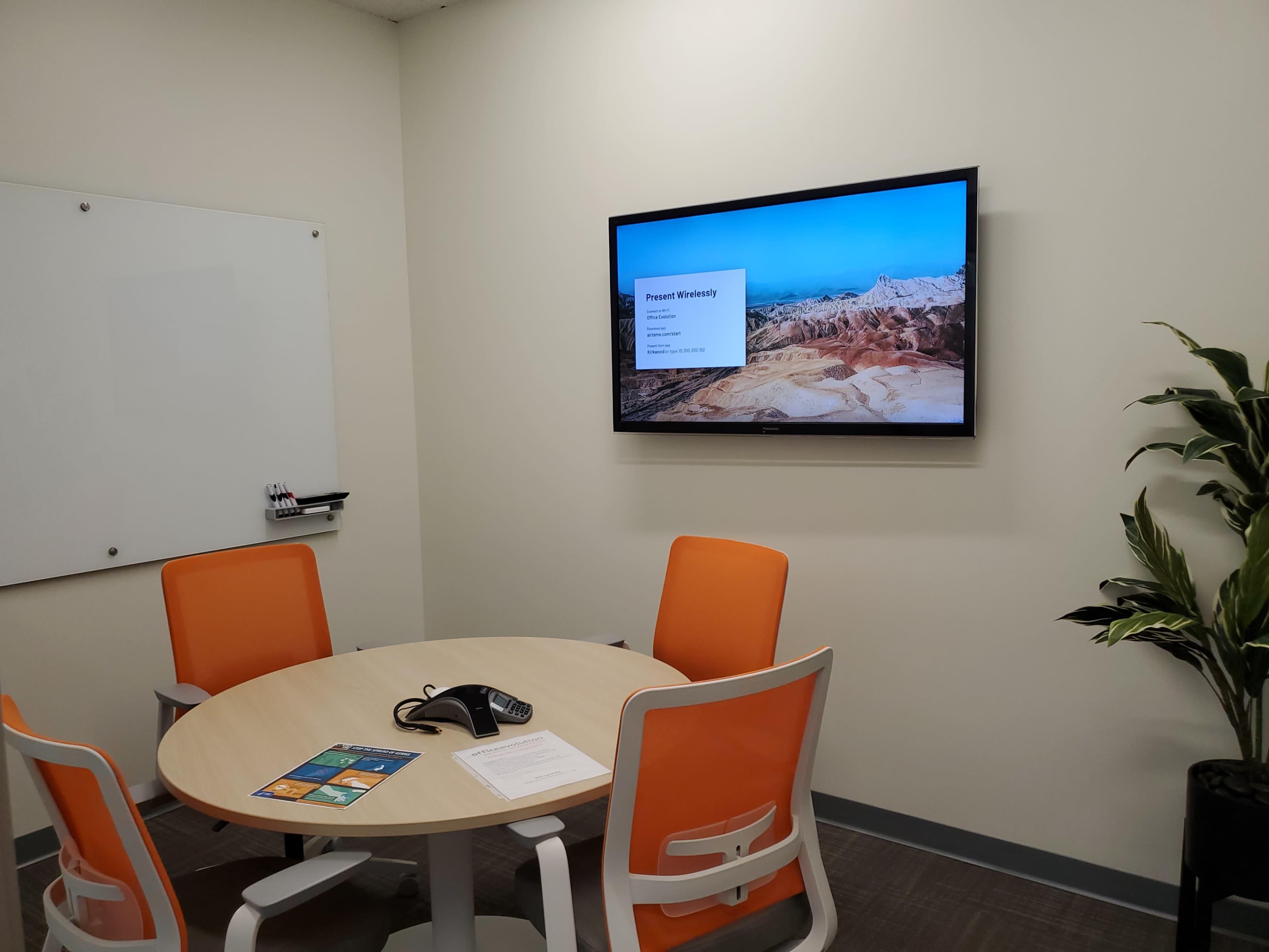 A small conference room features a round table with four orange chairs, a telephone, and a wall-mounted screen displaying a presentation.