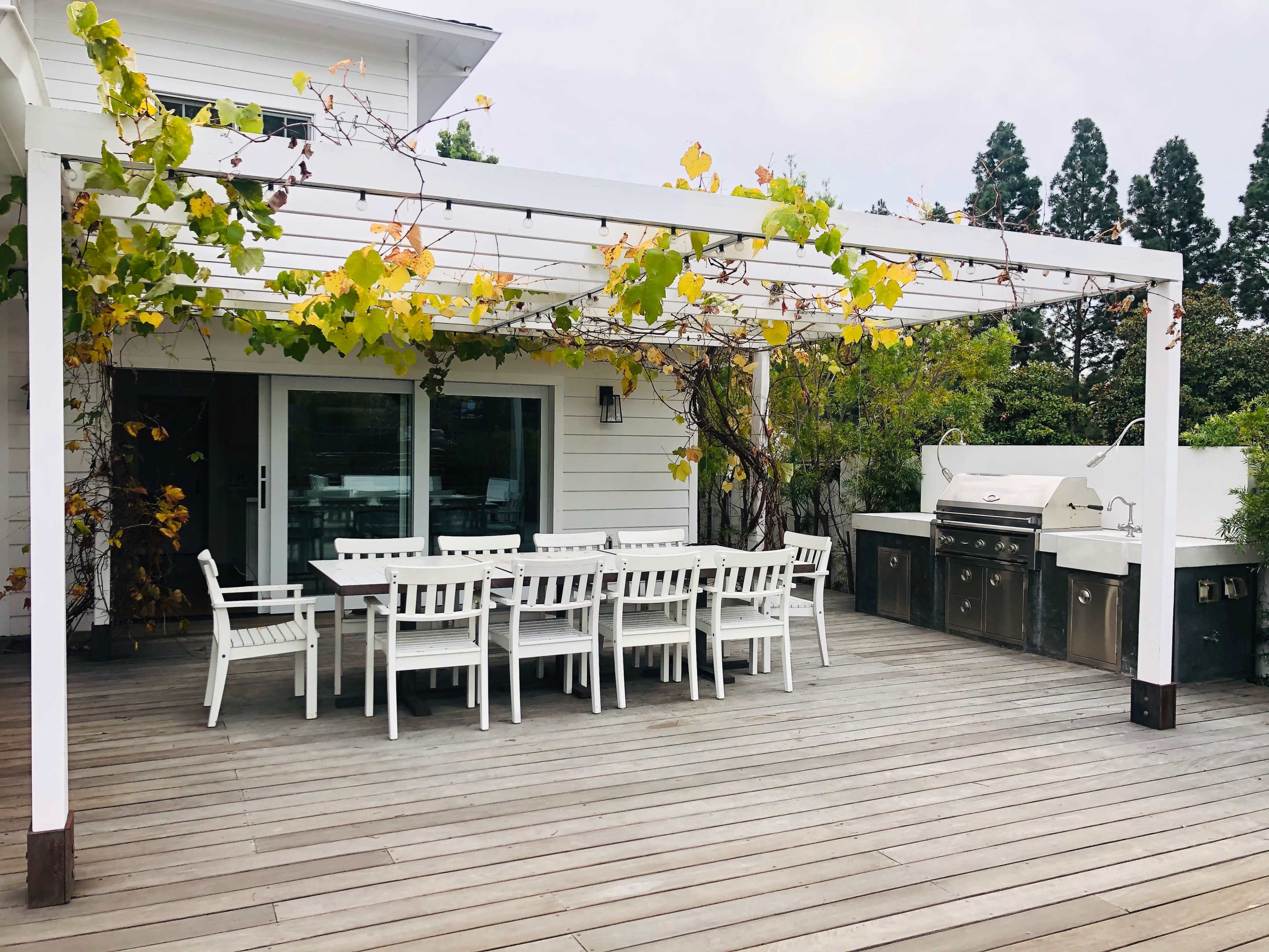 A spacious outdoor dining area with a large table and chairs, covered by a pergola adorned with climbing vines, adjacent to a built-in barbecue grill.