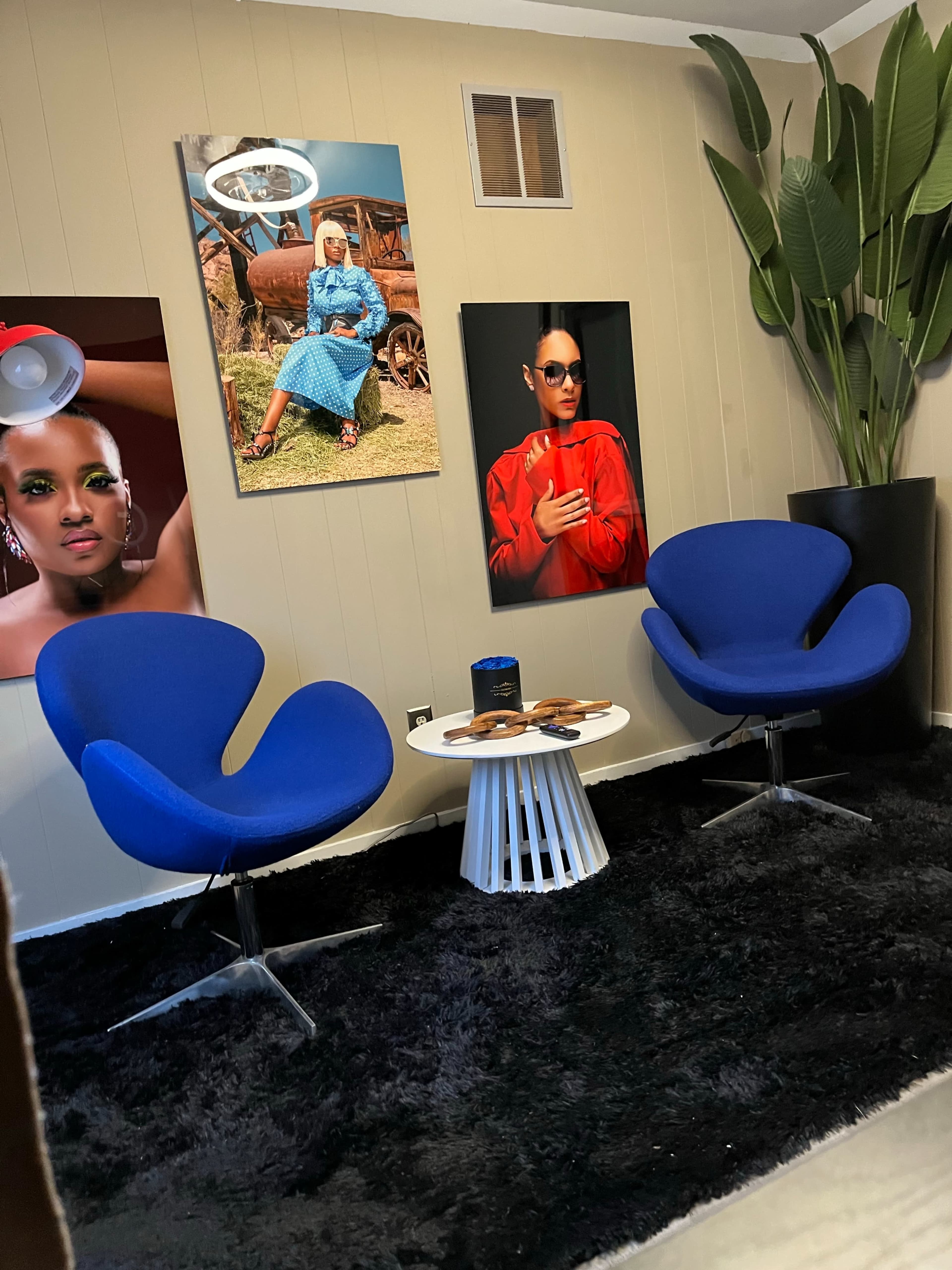 The image shows a cozy corner of a room with two blue chairs, a small white table, and large decorative plants, accompanied by colorful wall art featuring portraits.