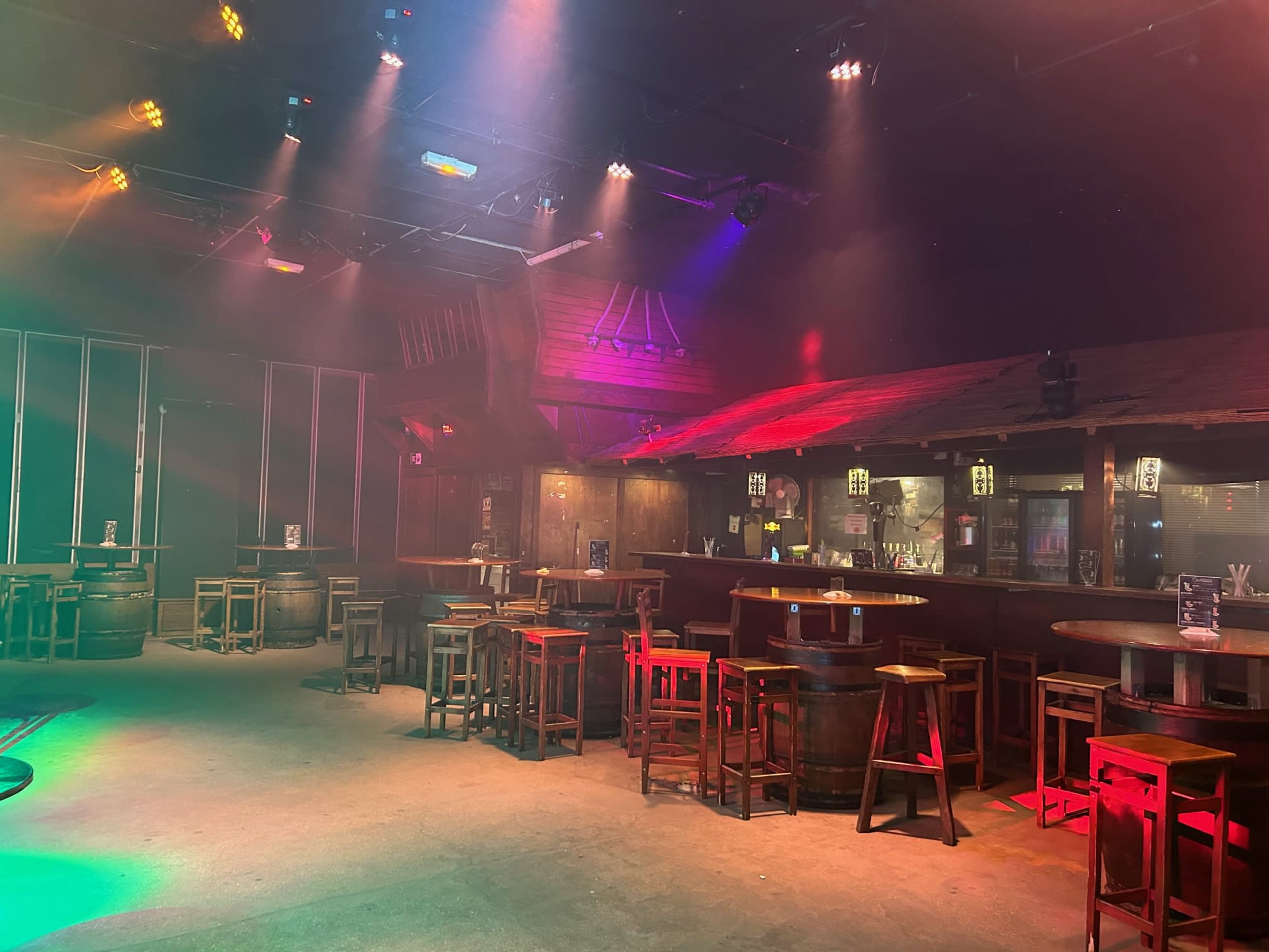 An empty bar space with wooden tables and chairs, illuminated by colorful stage lights.