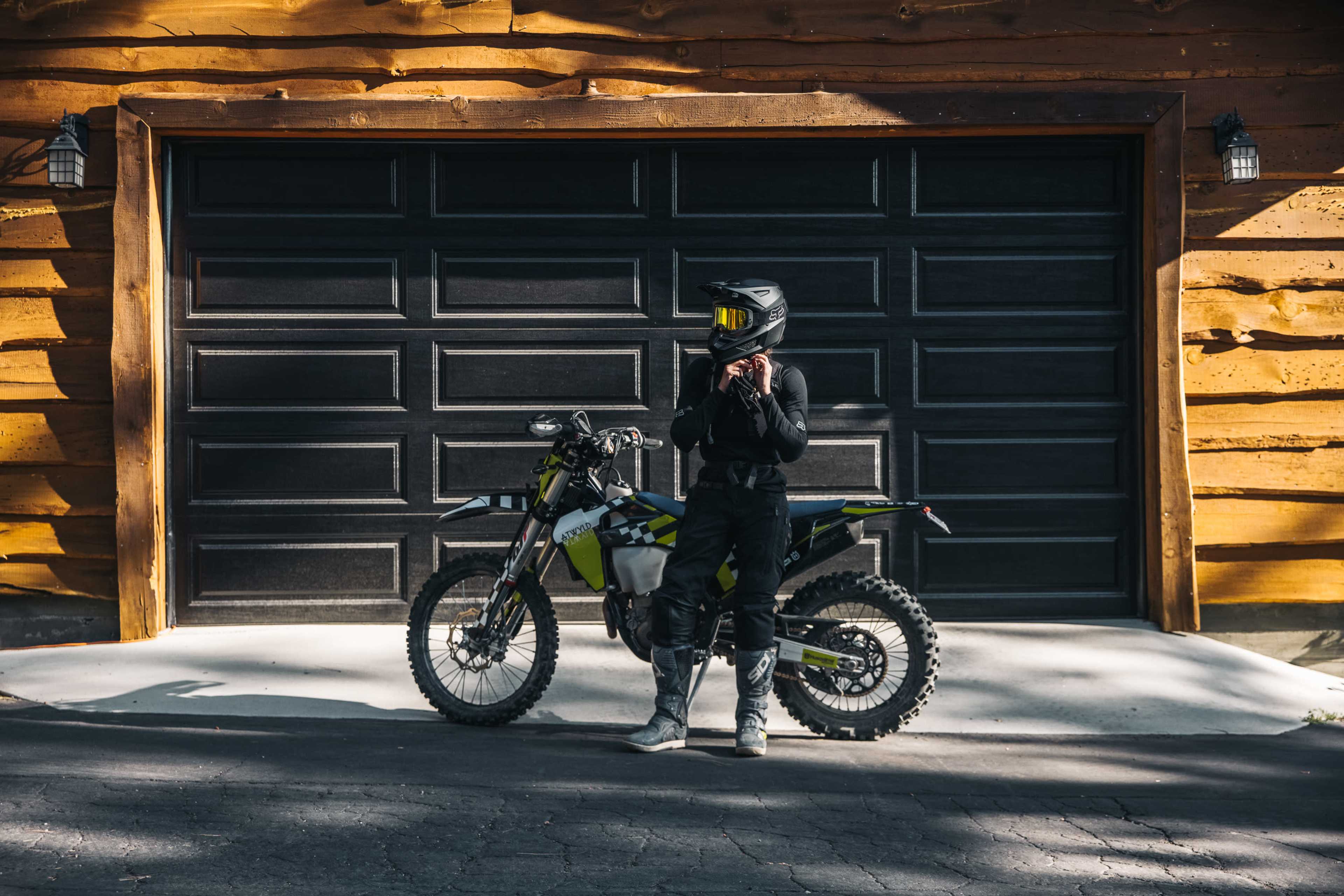A person in a black outfit and helmet stands beside a dirt bike in front of a wooden garage door.