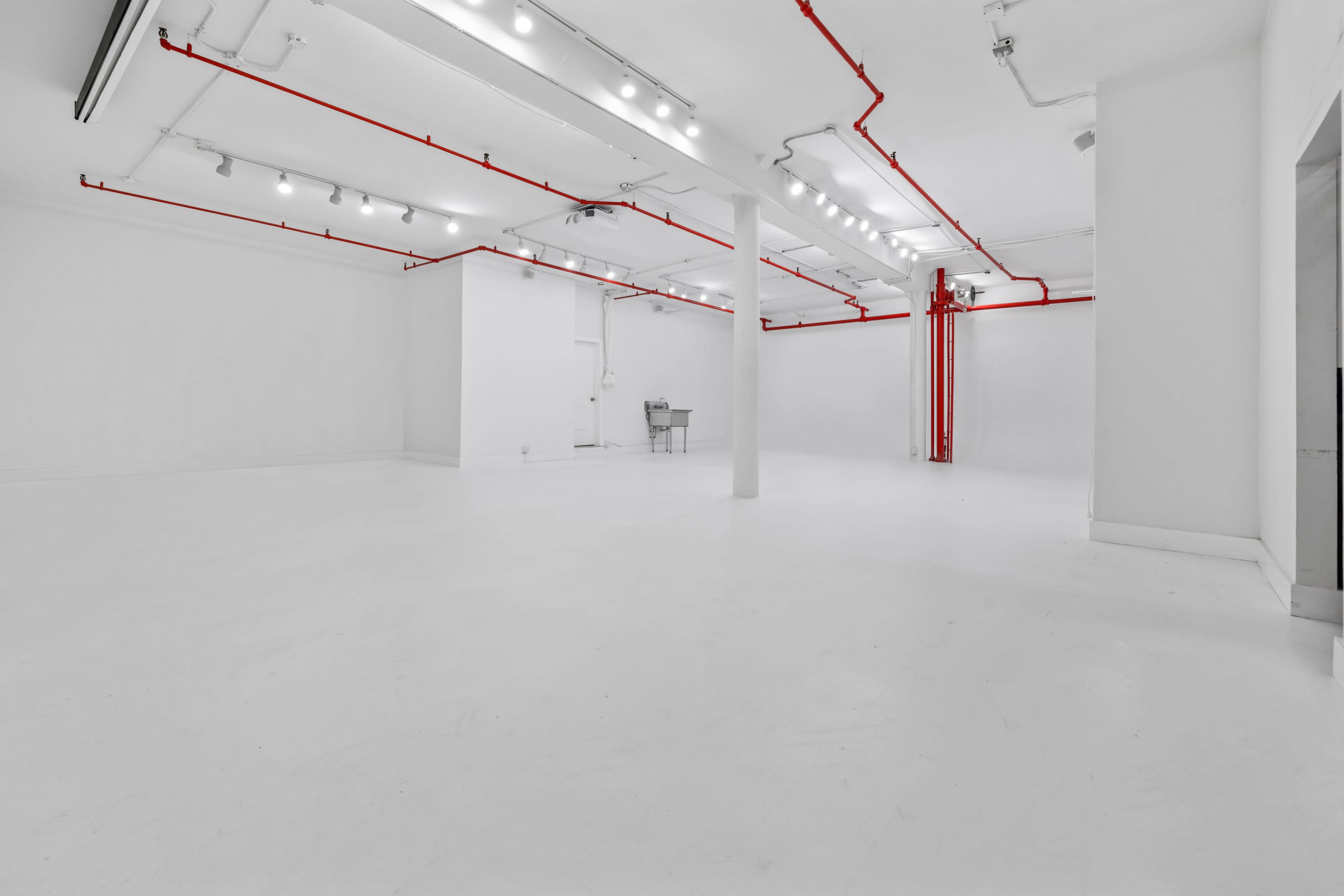 The image shows an empty room with white walls and a white floor, featuring red pipes along the ceiling and a single metal cart in the background.