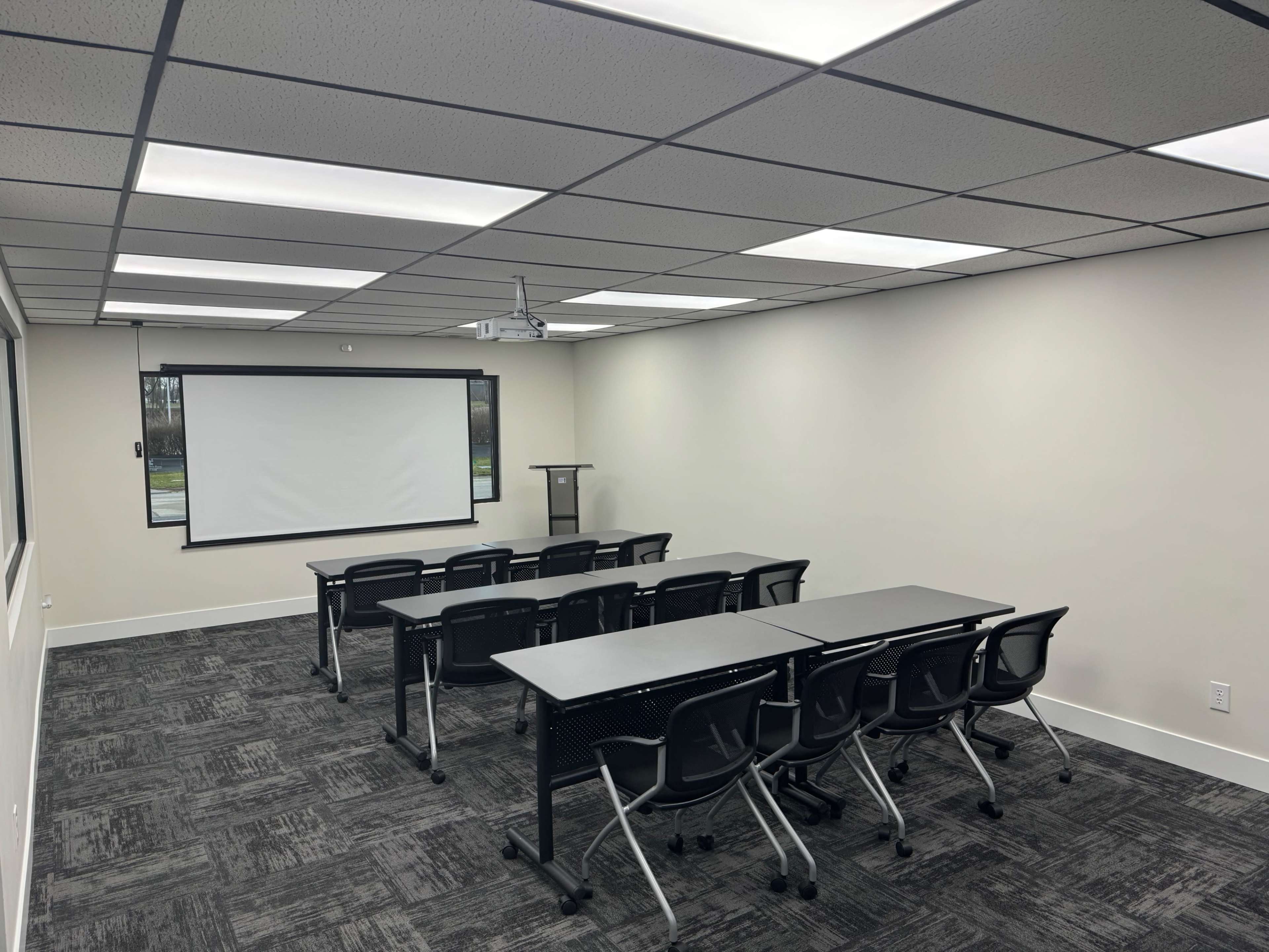 A modern, empty classroom or conference room arranged with several tables and chairs facing a projector screen at the front.