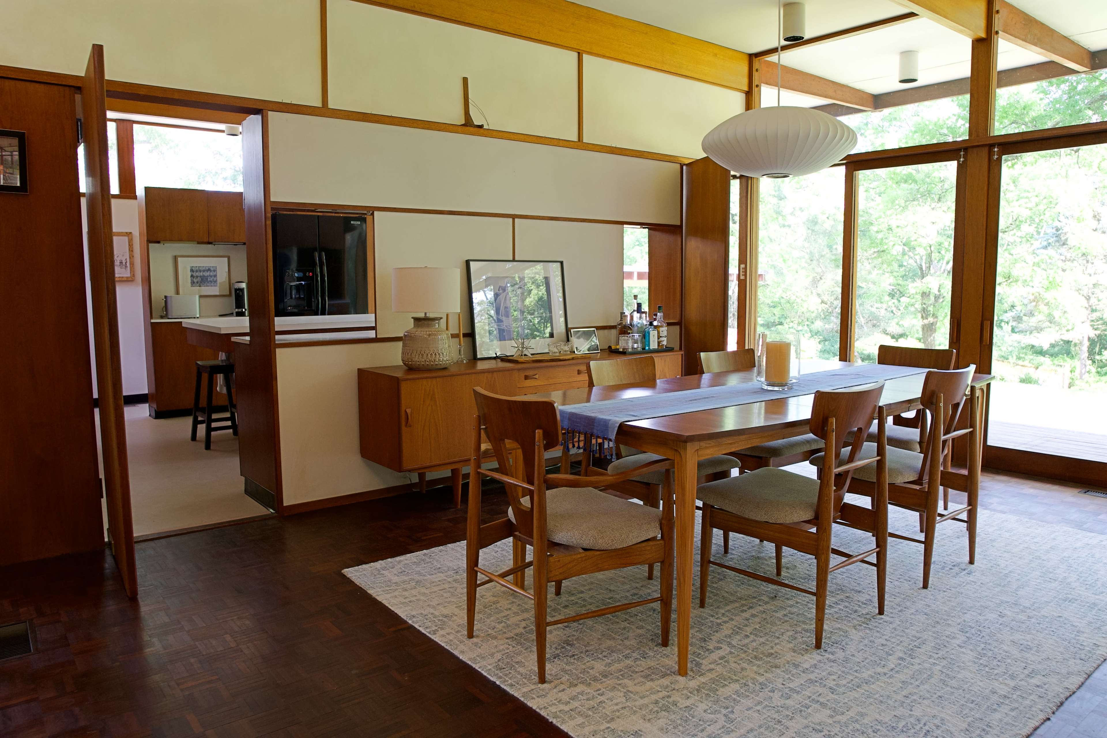A modern dining area features a wooden table surrounded by six chairs, with a sideboard and a large window overlooking greenery.