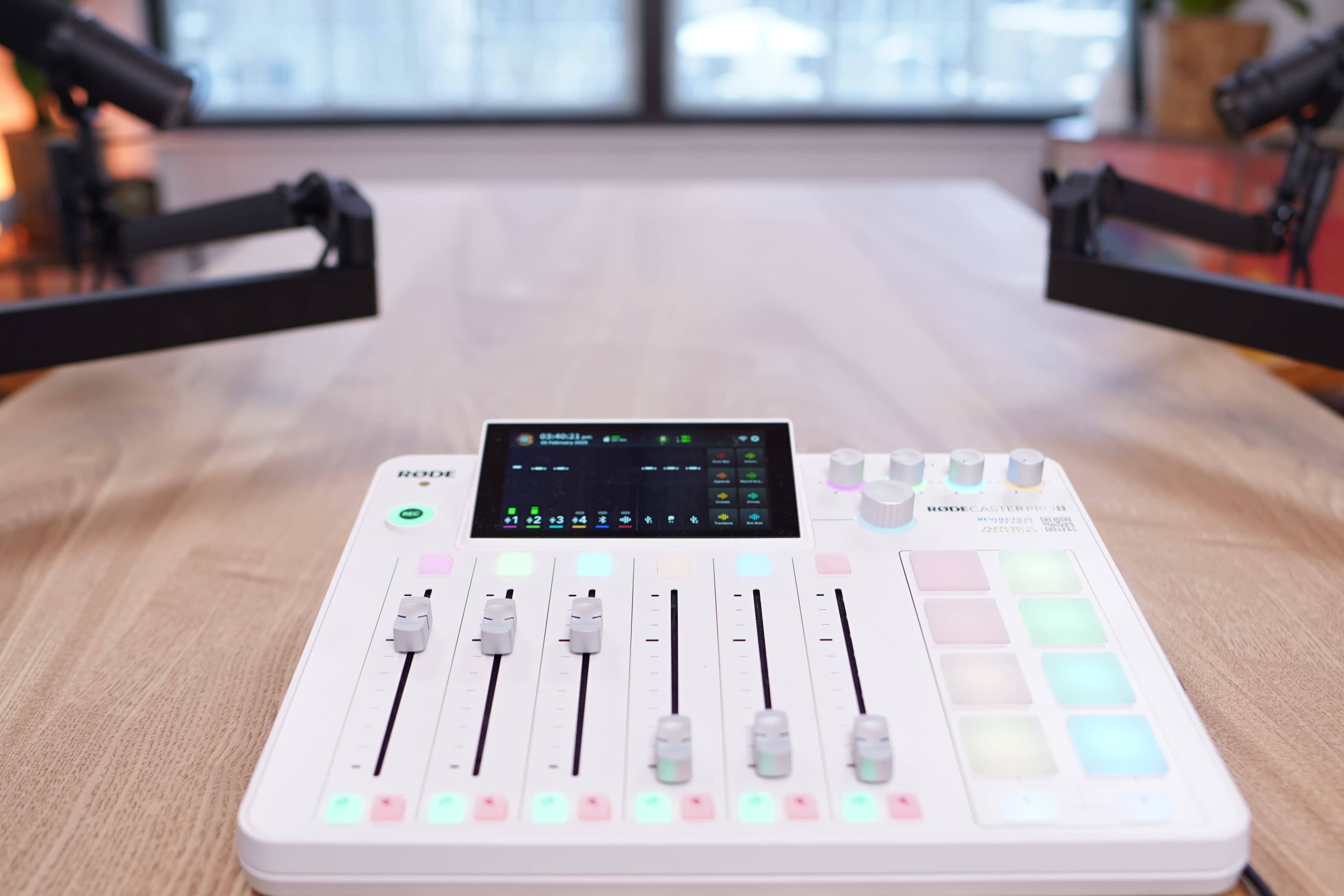 A fader control device with sliders, buttons, and a touchscreen sits on a wooden table between two microphones.