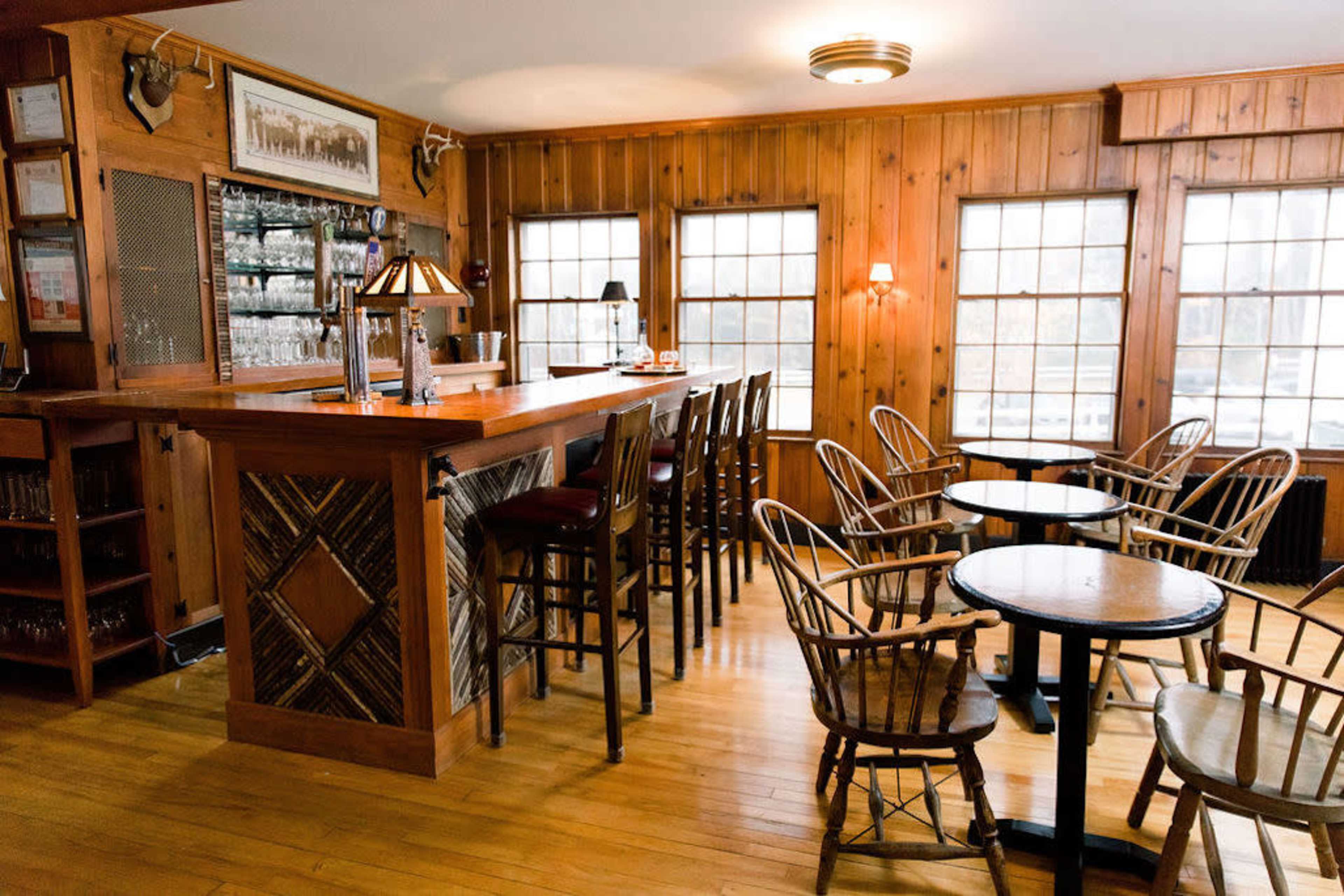 The interior of a rustic bar features wooden paneling, a polished bar with stools, and small round tables surrounded by wooden chairs.