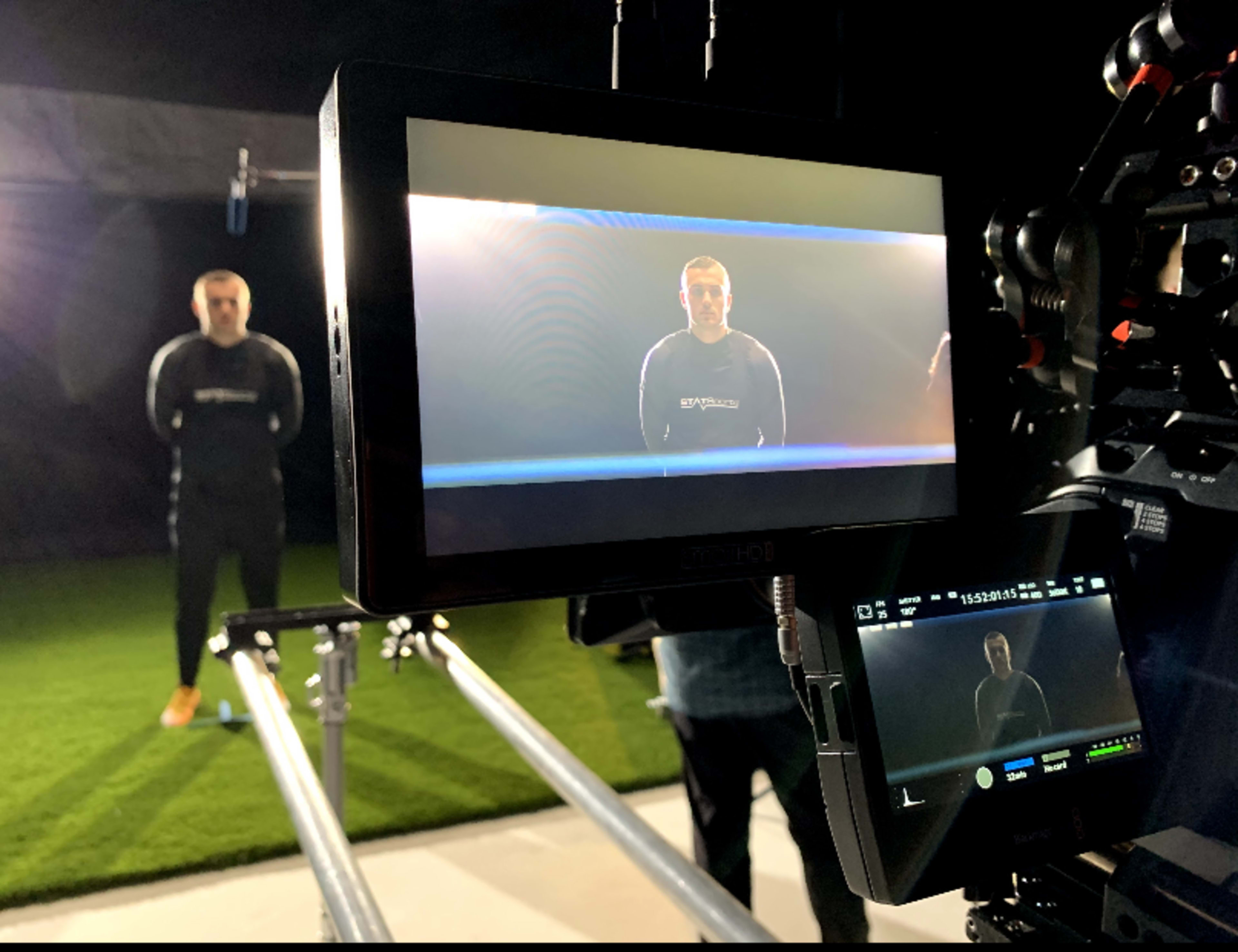 A person stands in front of a camera setup on a grassy surface, while the camera displays a focused view of the subject on its monitor.