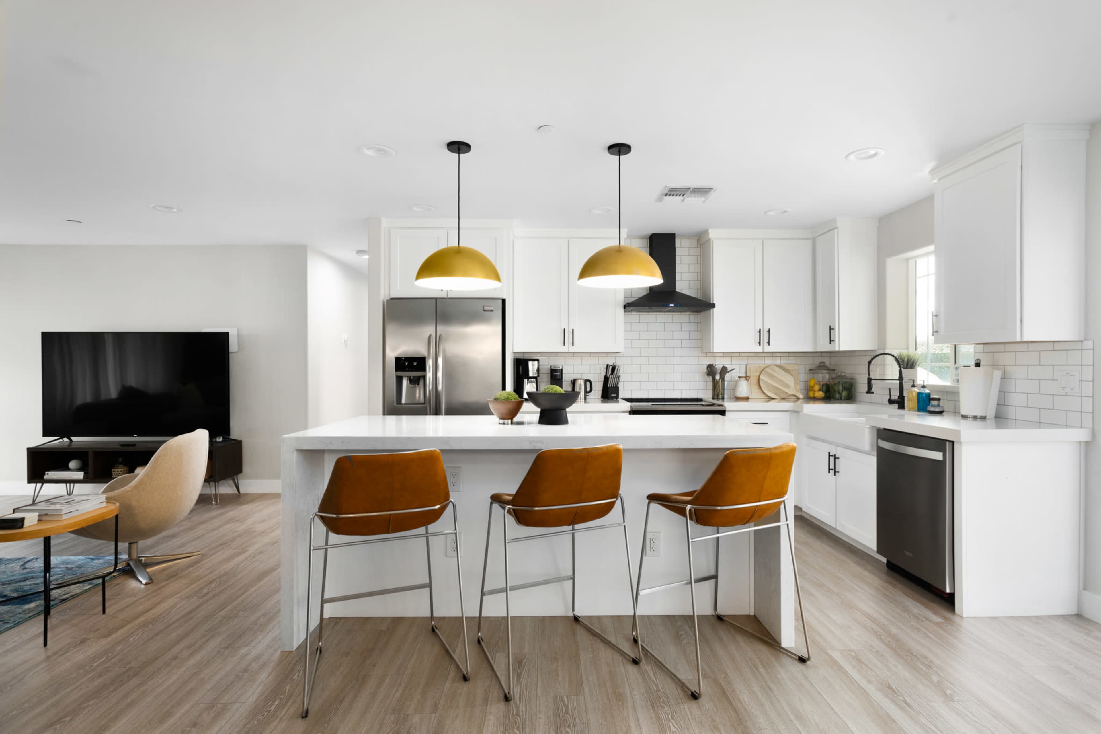A modern kitchen features white cabinetry, a large island with three bar stools, and two pendant lights above, with a living area visible in the background.