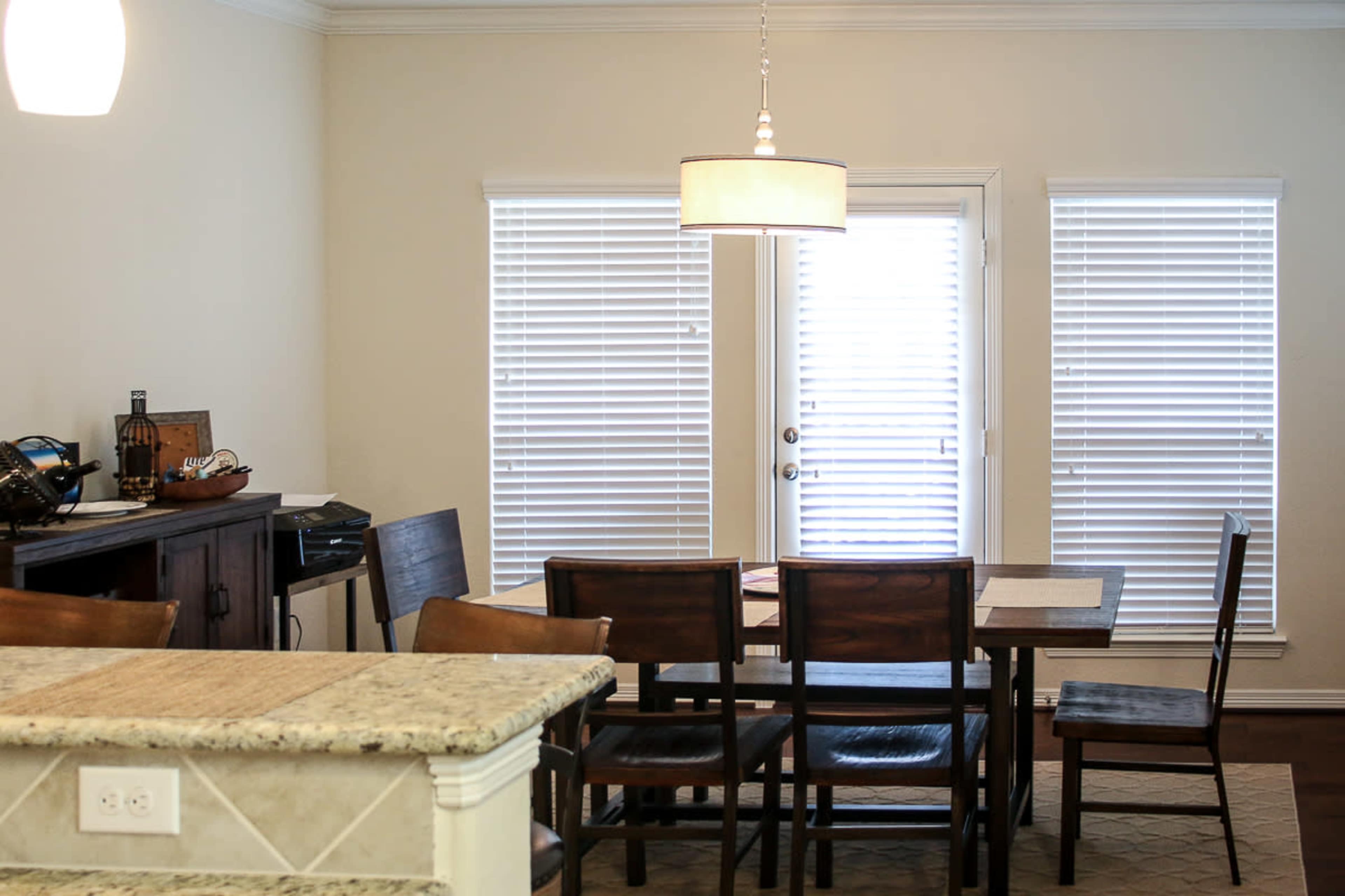 The image shows a dining area with a wooden table and chairs, a light fixture overhead, and windows with closed blinds.