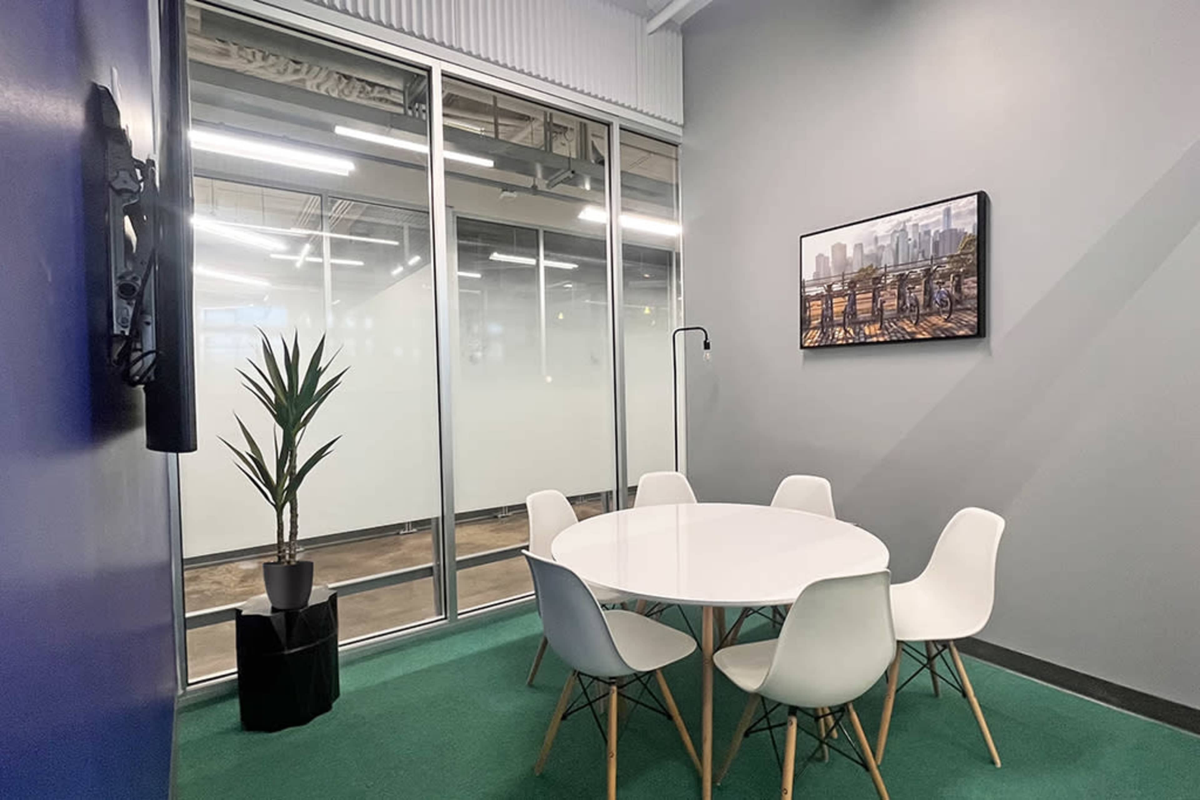 A small conference room features a round white table surrounded by six chairs, a potted plant, and a large wall-mounted television.