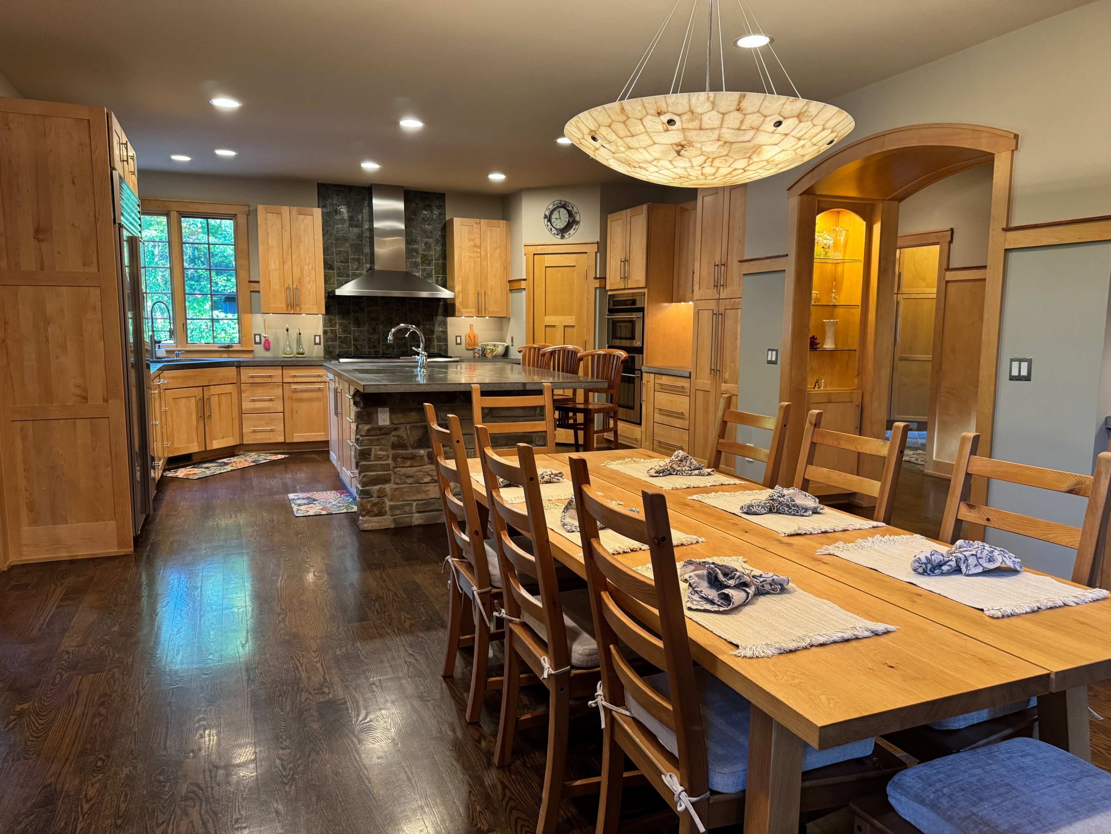 The image shows a spacious kitchen and dining area with wooden cabinetry, a large island, and a long dining table set with placemats.