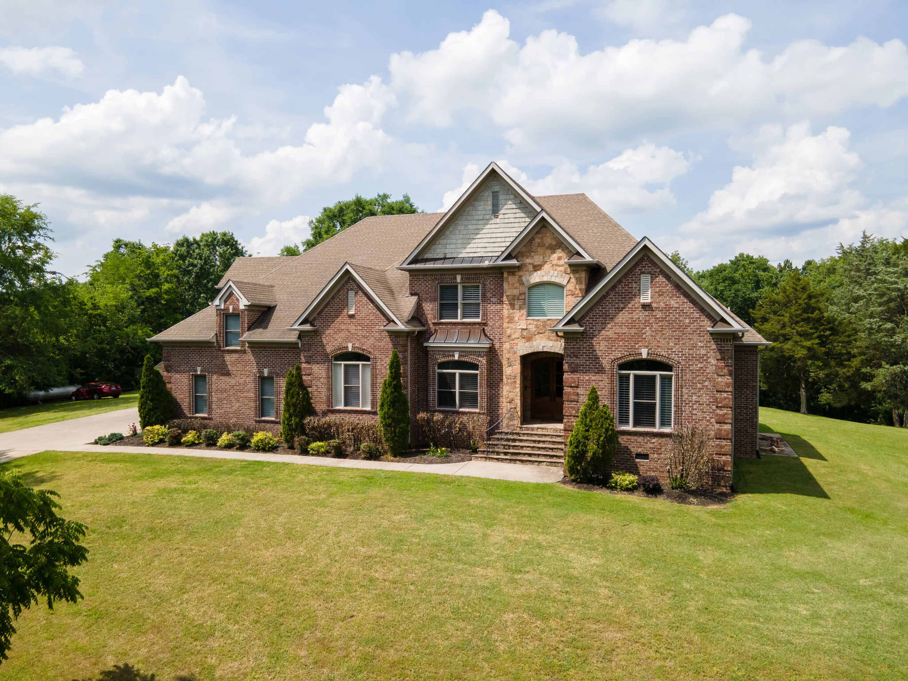 A large brick house with a steeply pitched roof is set on a spacious green lawn, surrounded by neatly trimmed bushes and trees.