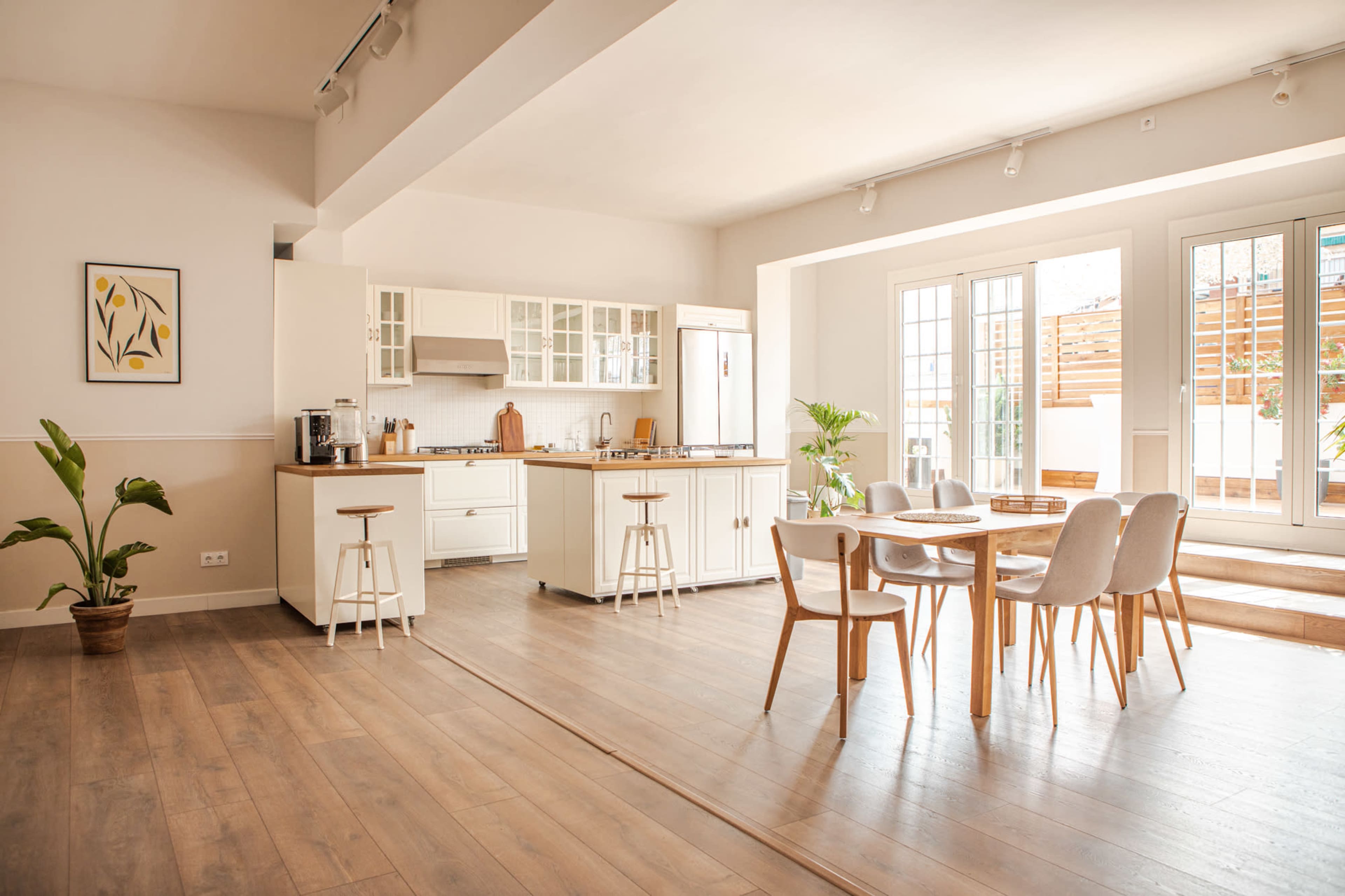 A bright kitchen features white cabinetry, a central island with stools, and a dining table surrounded by chairs, with large windows allowing natural light to illuminate the space.