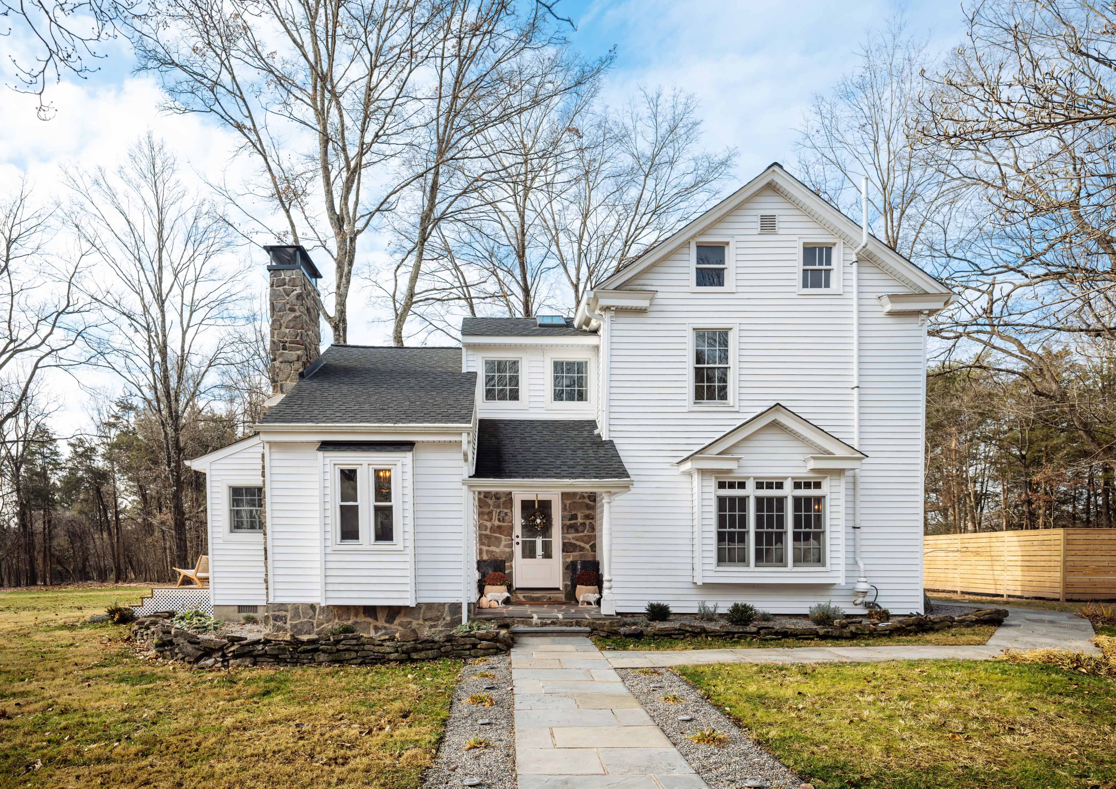 A white, two-story house with a stone chimney and a well-maintained yard is set among bare trees.