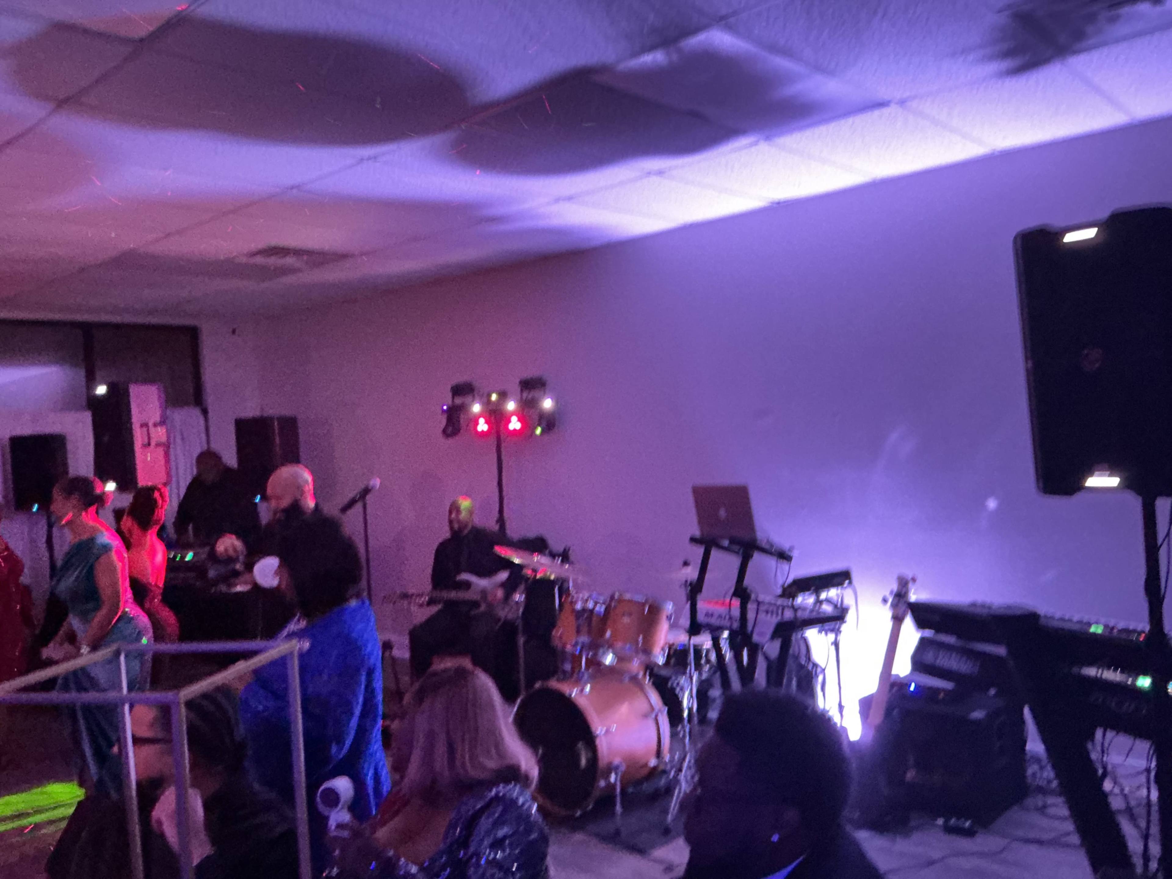 The image shows a lively indoor event with a live band setup, including a drum kit and electronic instruments, while guests dance and socialize in the background.