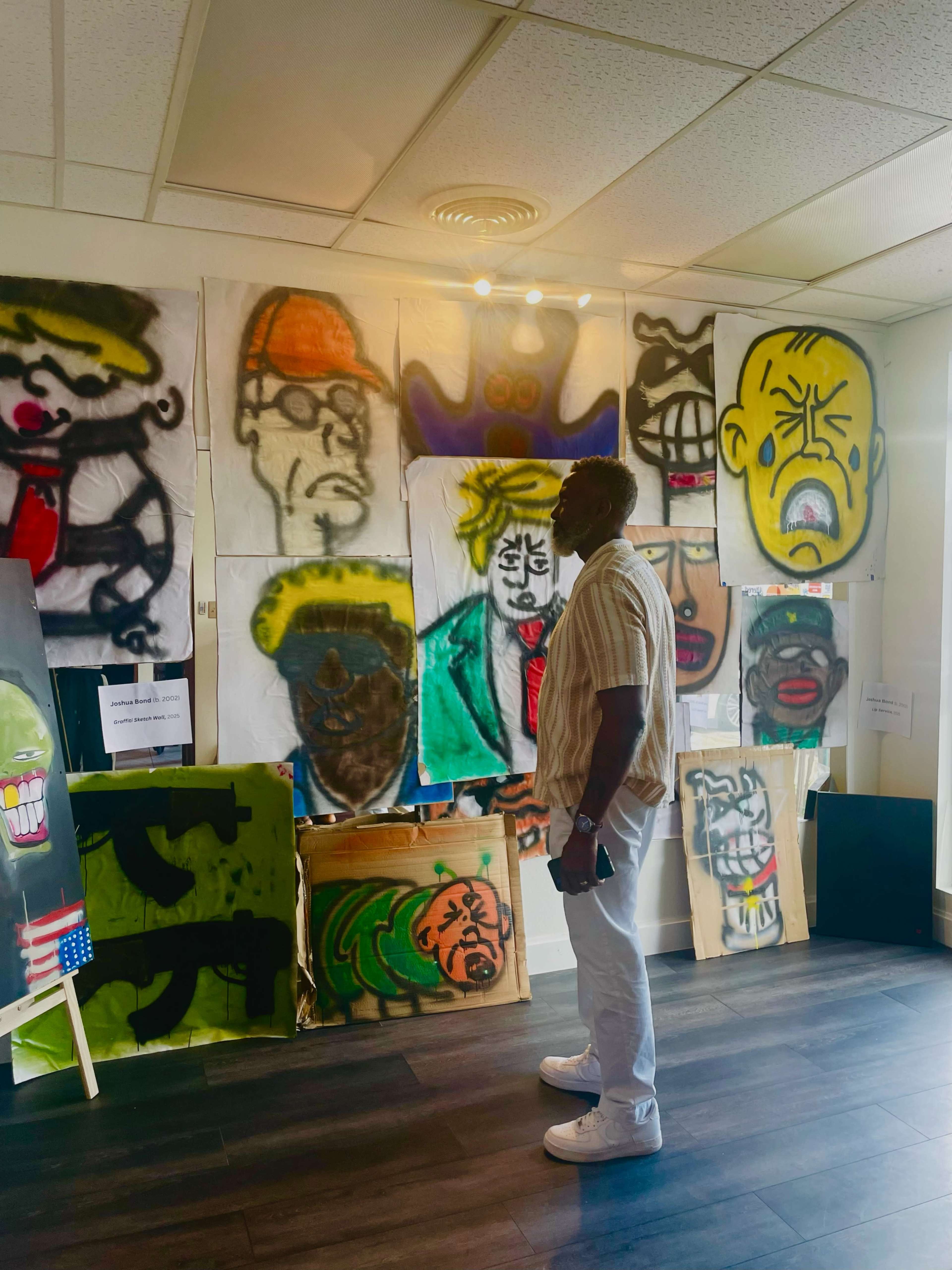 A man stands in front of a wall covered with various colorful, oversized spray-painted portraits.