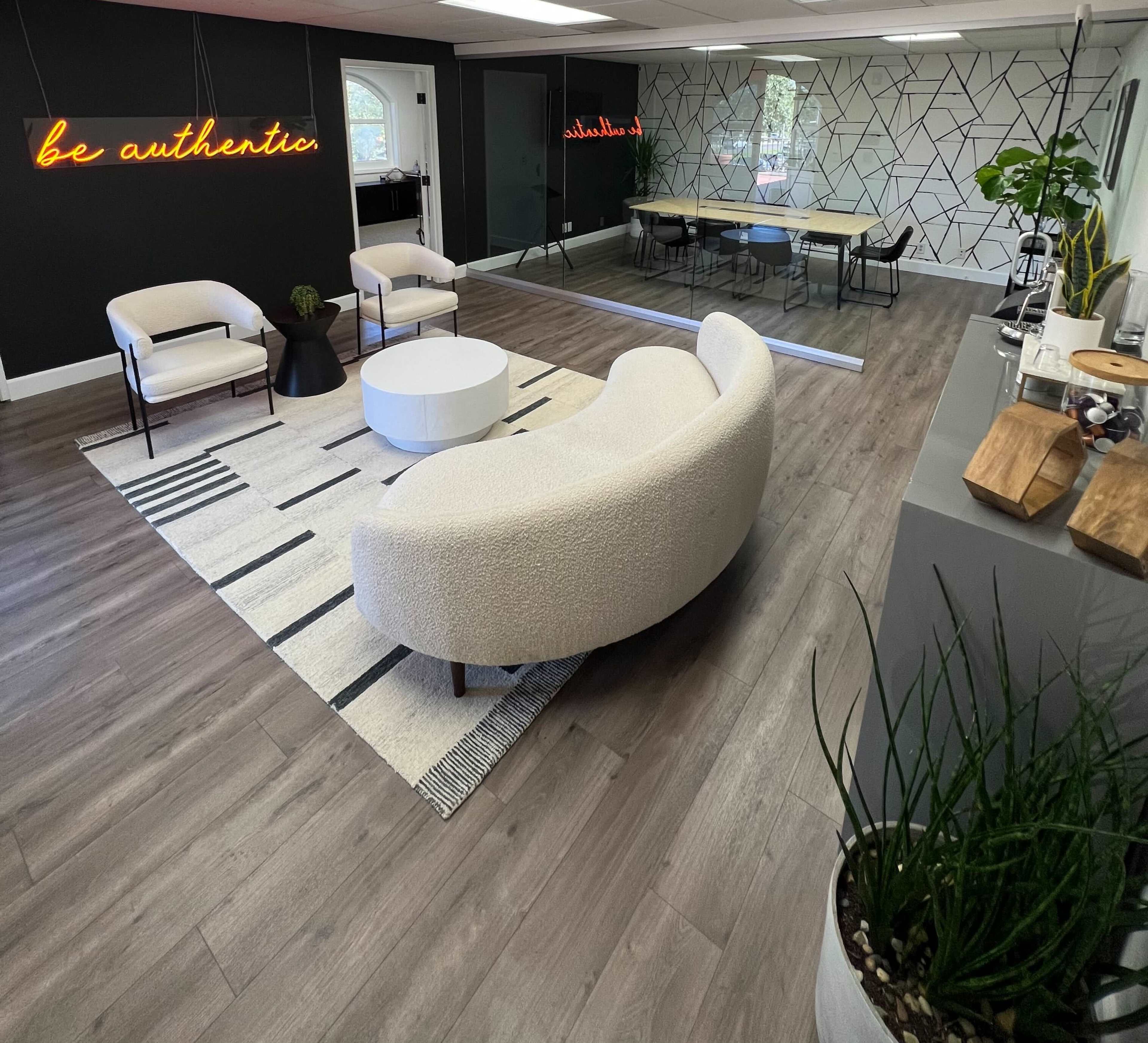 A modern office space featuring a curved white sofa, a round coffee table, and a meeting area with a long table in the background, accented by a neon sign that reads "be authentic."