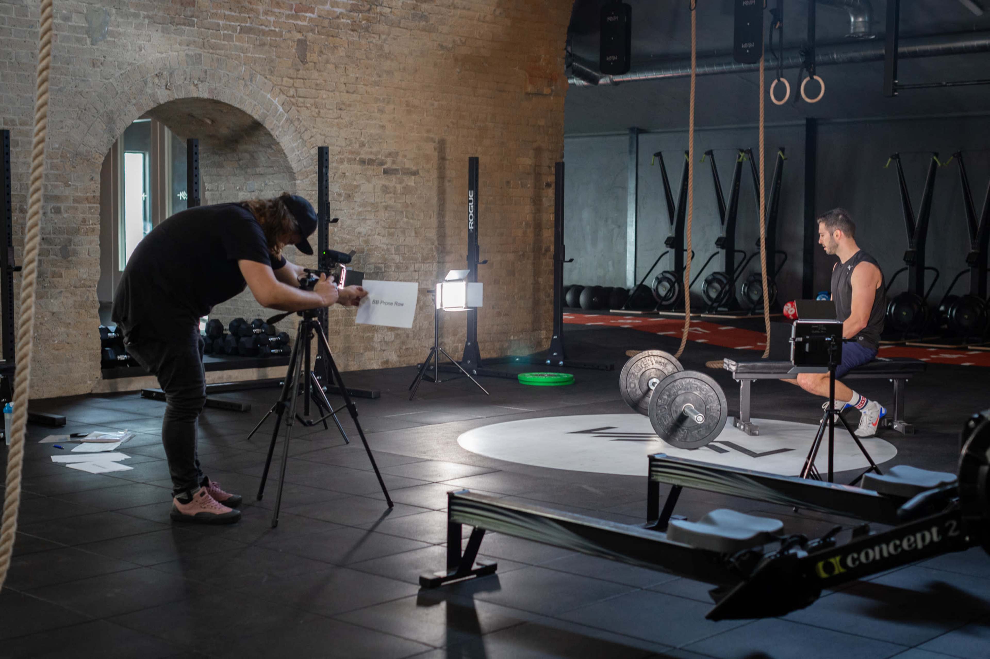 A fitness trainer is photographed while preparing to lift weights in a gym, with cameras set up for a video shoot.