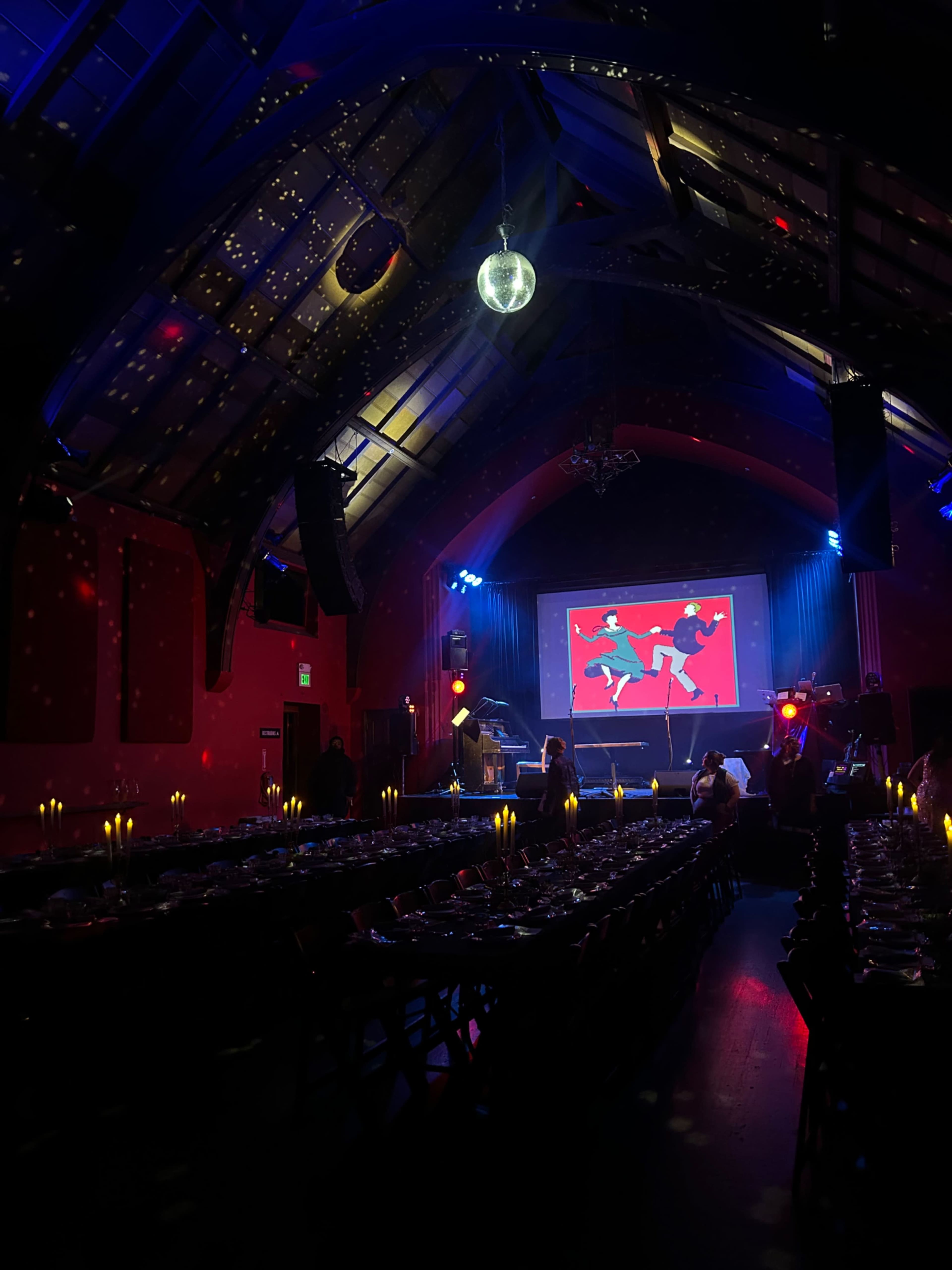 A dimly lit banquet hall features long tables set for dinner, with a large screen displaying a colorful graphic at the front.