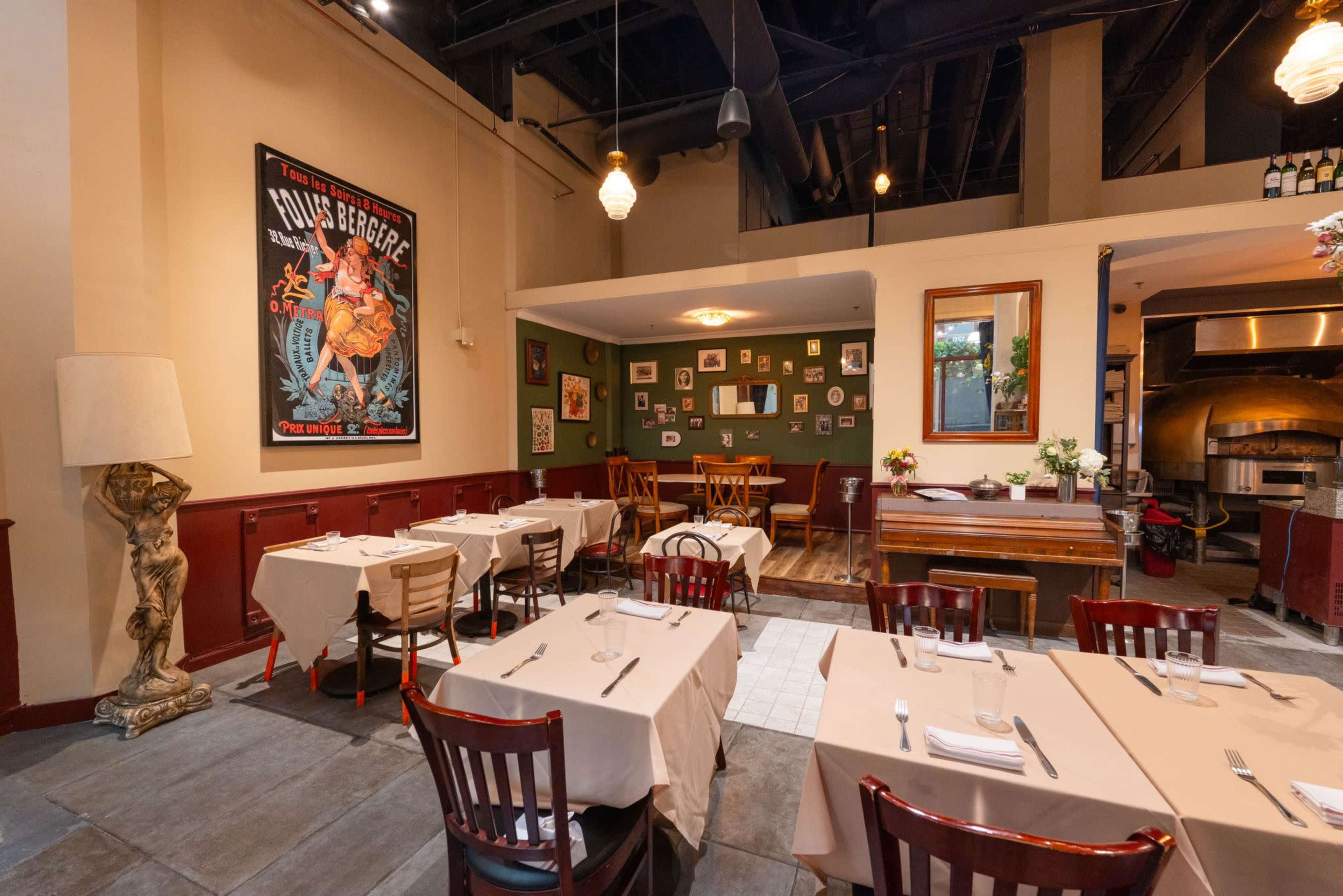 The interior of a restaurant features wooden chairs, beige tablecloths, and decorative wall art, with a wood-fired oven visible in the background.