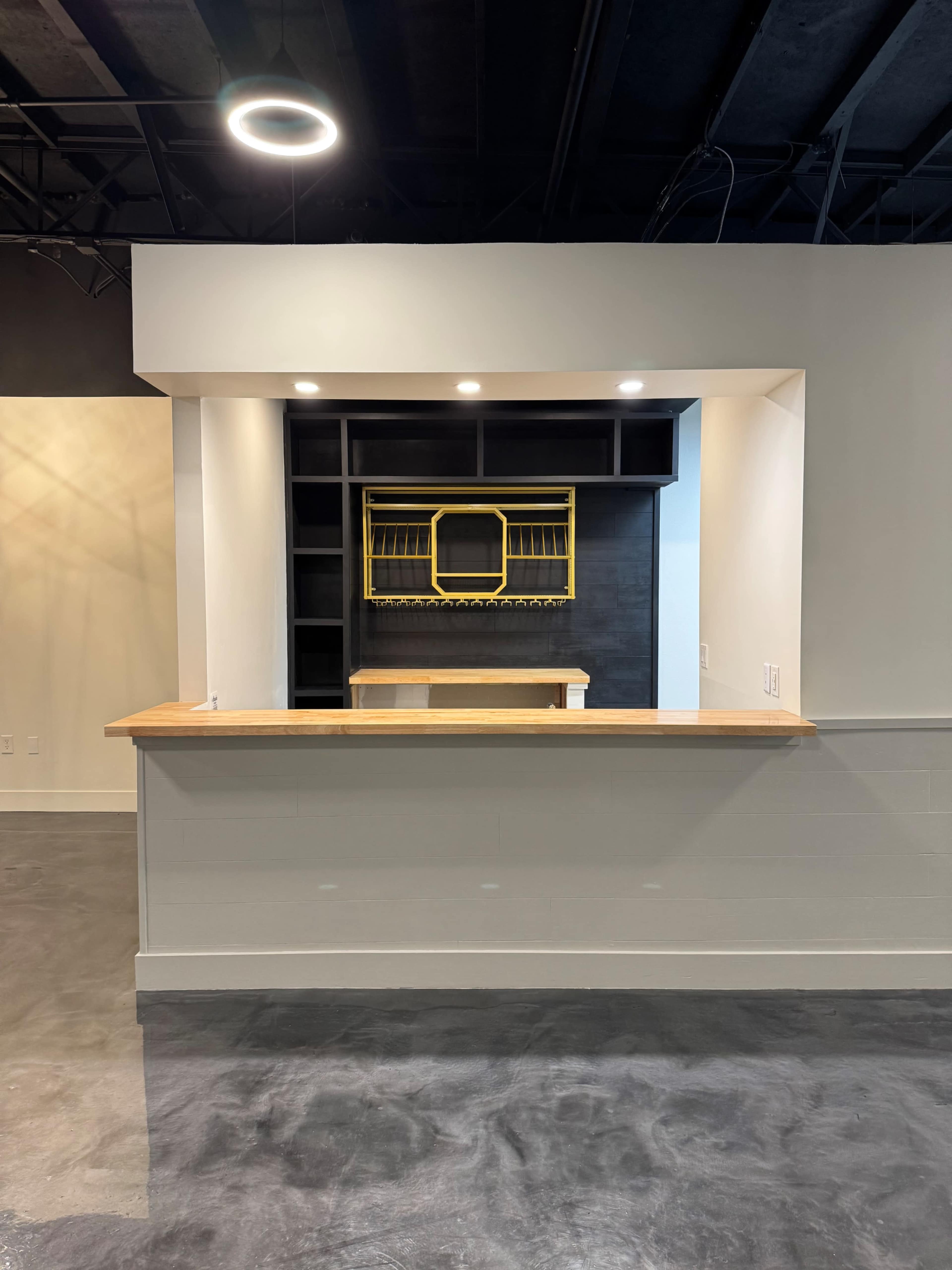 The image shows a minimalist reception area featuring a wooden counter and a wall shelf with a yellow frame, illuminated by recessed lighting.