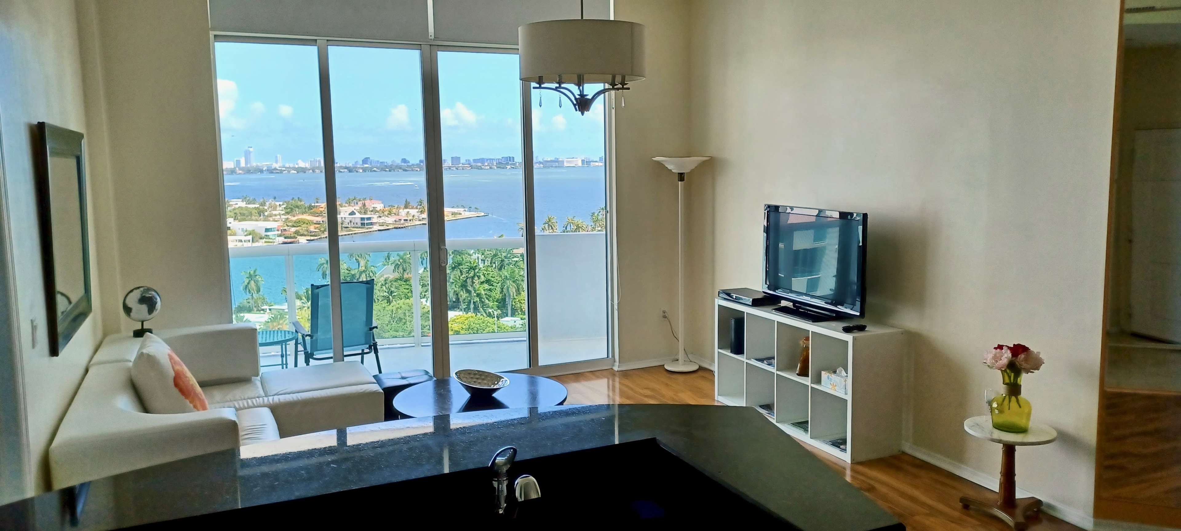 A modern living room with large windows overlooking a bay, featuring a couch, a coffee table, and a television.
